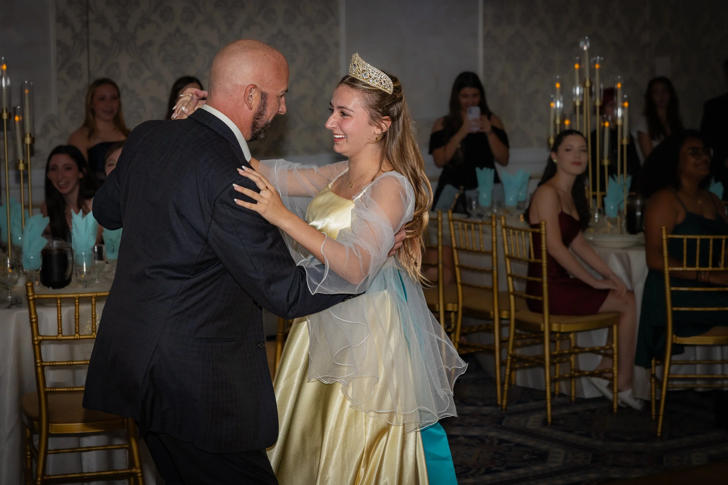 A man and a young woman dancing together at a celebration, with the woman wearing a tiara and a dress resembling a princess costume. Several people are seated at tables in the background, some taking photos and smiling.