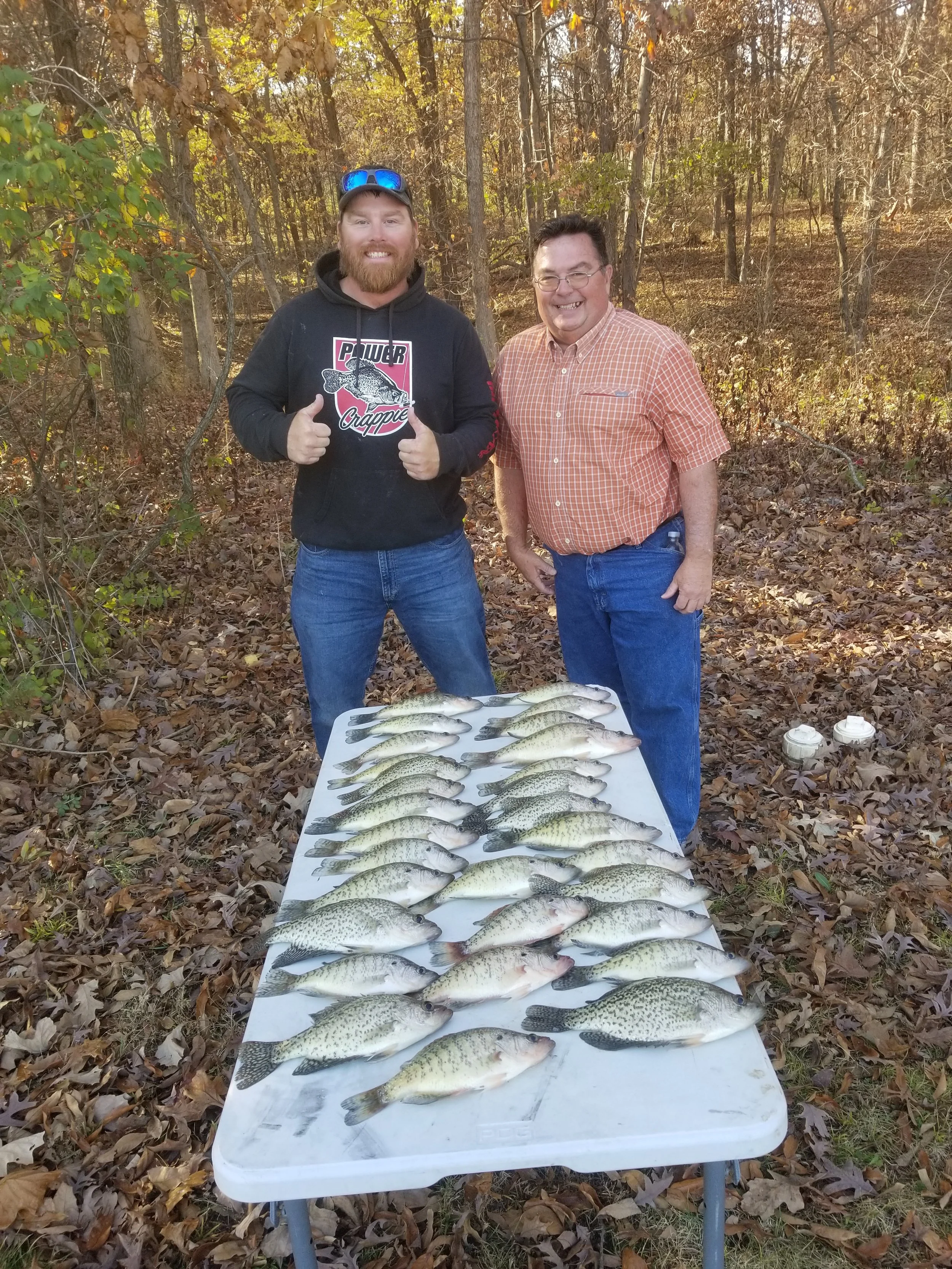 Two men standing outdoors in a wooded area with fallen leaves, smiling for the camera. One man is giving a thumbs-up, wearing a black hoodie with a fish graphic. The other man is wearing a plaid button-up shirt. They are standing behind a table covered with a white cloth, displaying a large catch of fish, all lined up in rows.