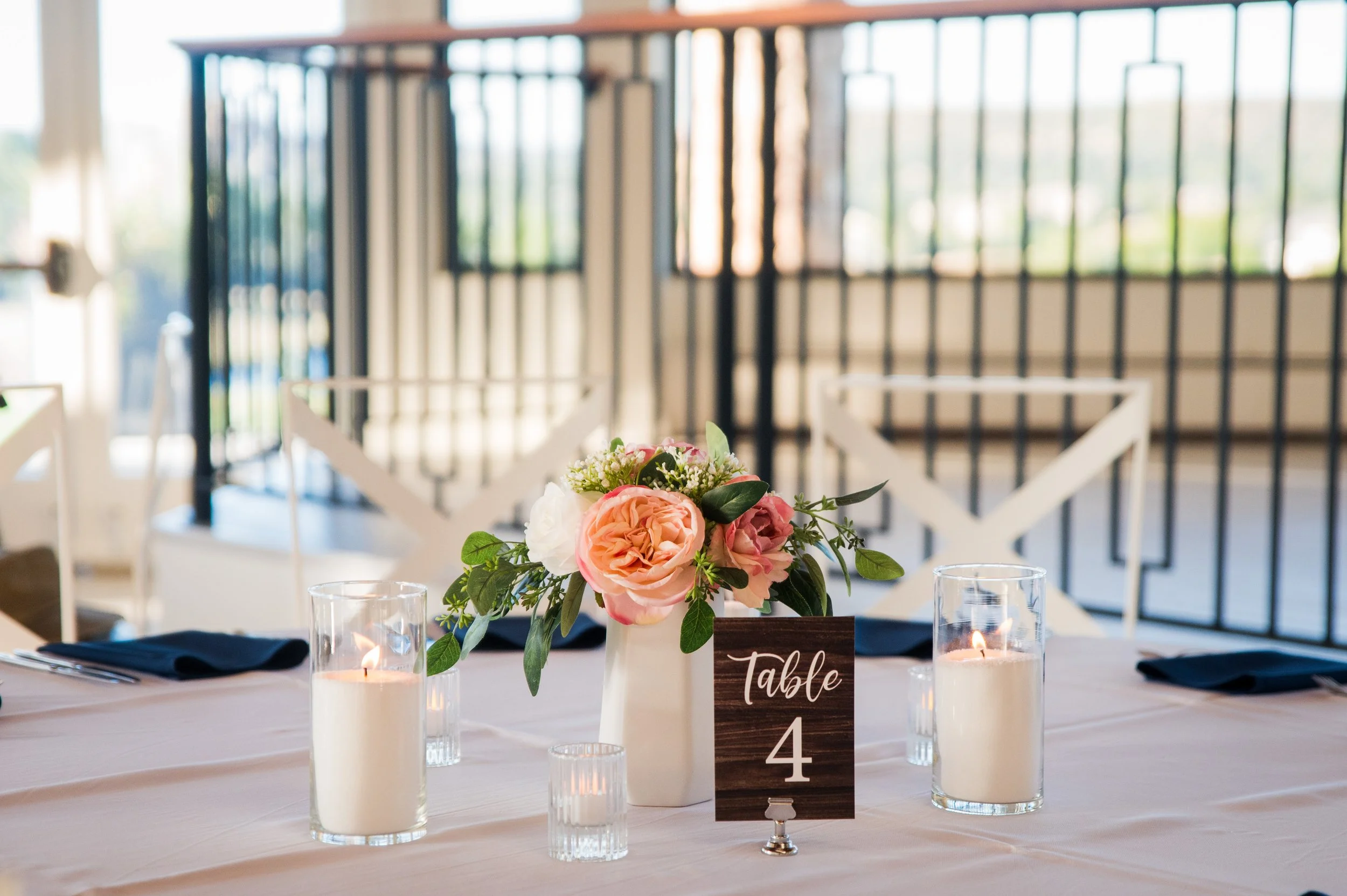 Elegant dining table centerpiece featuring pink and white flowers in a white vase, surrounded by candles and a table number card with the word 'Table' and the number 4, in a bright room with large windows.