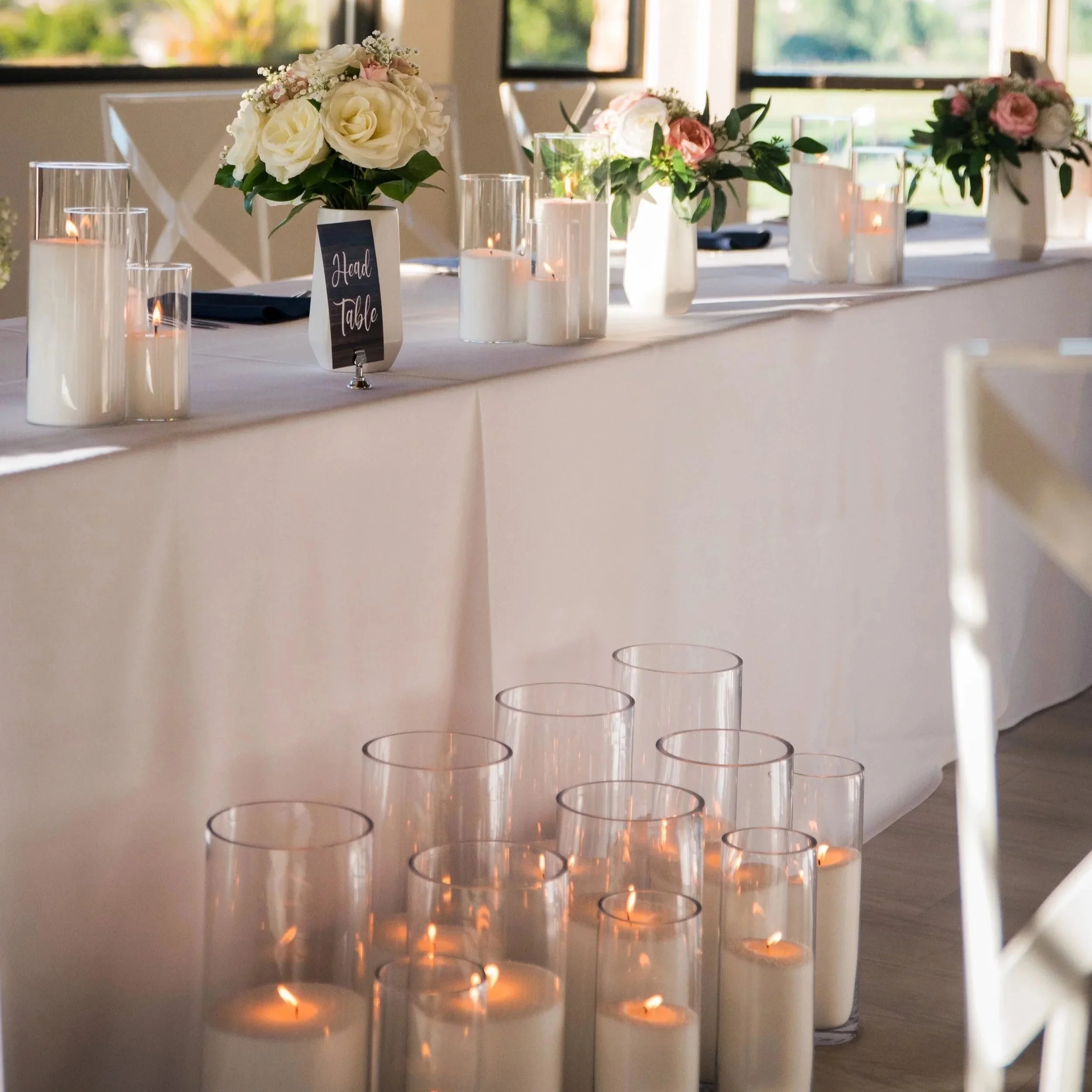A table decorated with white and pink flowers in white vases, surrounded by lit candles in glass holders, with a sign indicating 'Head Table.' The table is positioned near large windows letting in natural light, creating a warm and elegant atmosphere