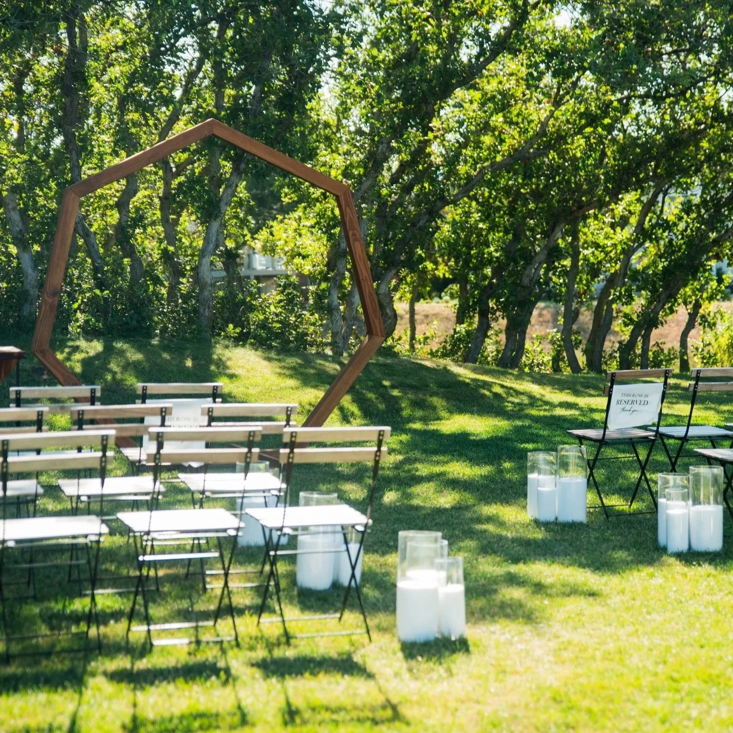 Outdoor wedding ceremony setup with rows of white chairs, a geometric wooden arch, and candles on the grass, surrounded by trees.