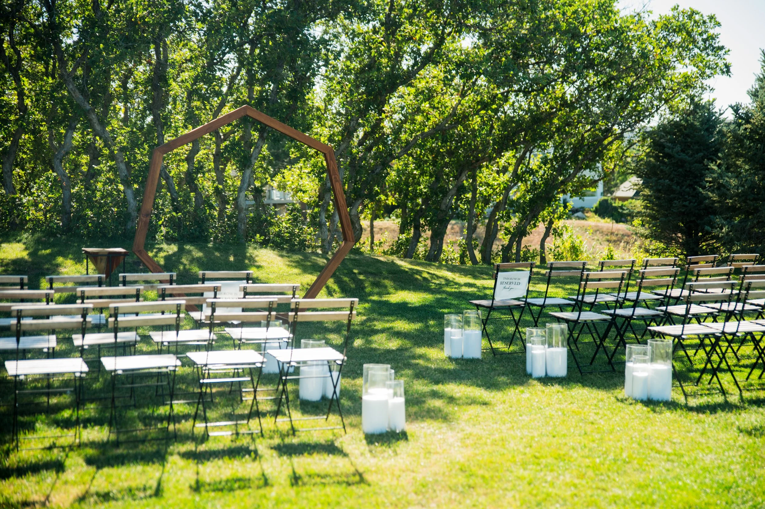 Outdoor wedding ceremony setup with rows of chairs, candles, and a wooden hexagon arch in a green park with trees.