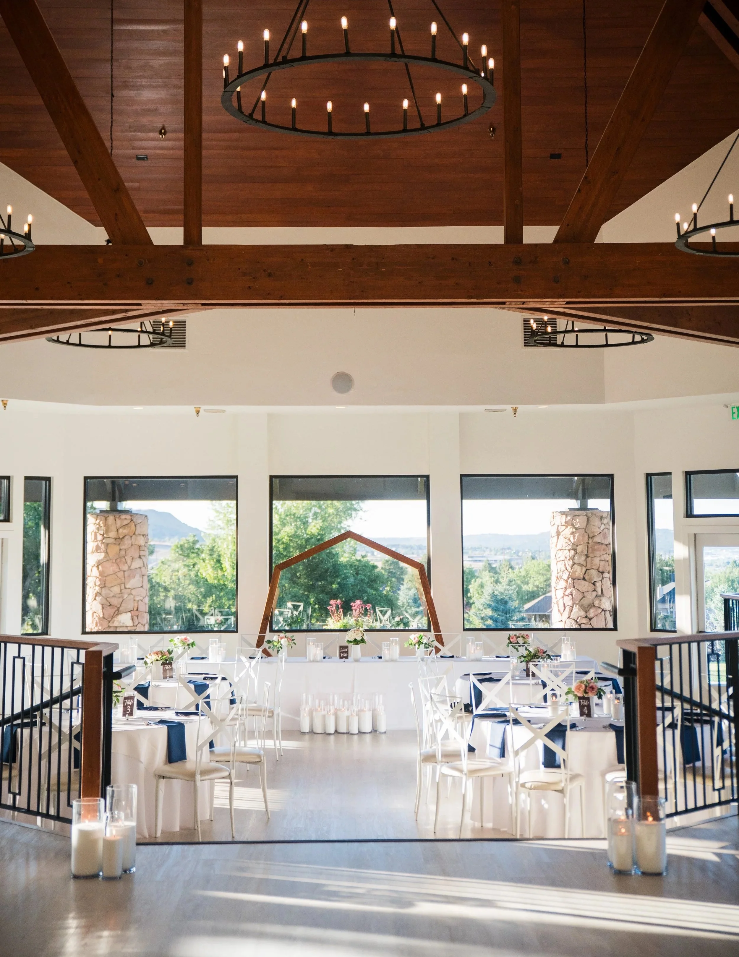 Indoor wedding reception area with white tables decorated with pink and white flowers, surrounded by white chairs, candles, and candles in glass holders. There are several large candles in glass holders on the floor in the foreground.