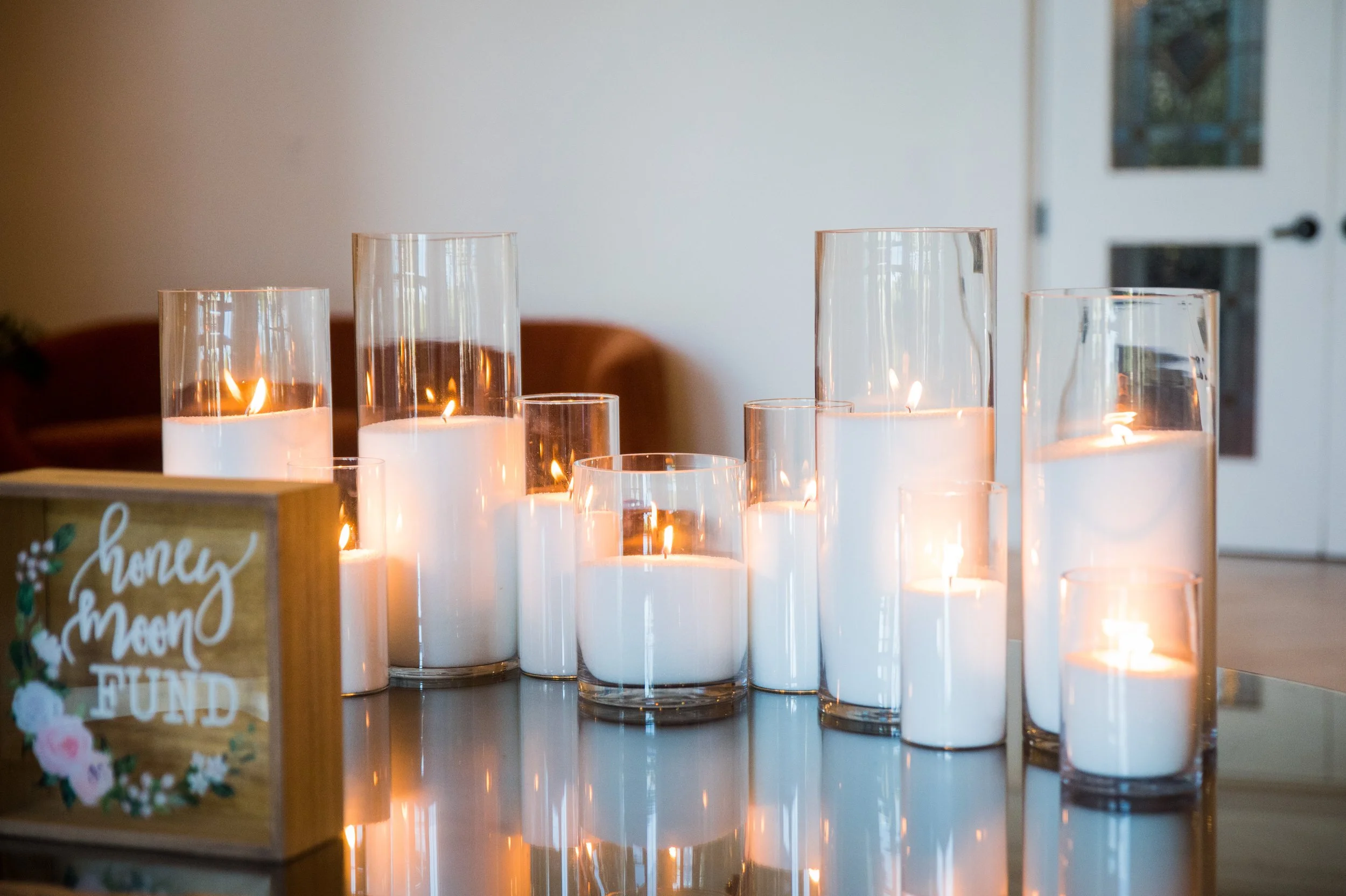 Several tall glass candle holders with lit candles inside on a table, with a blurred background including a wall and door.