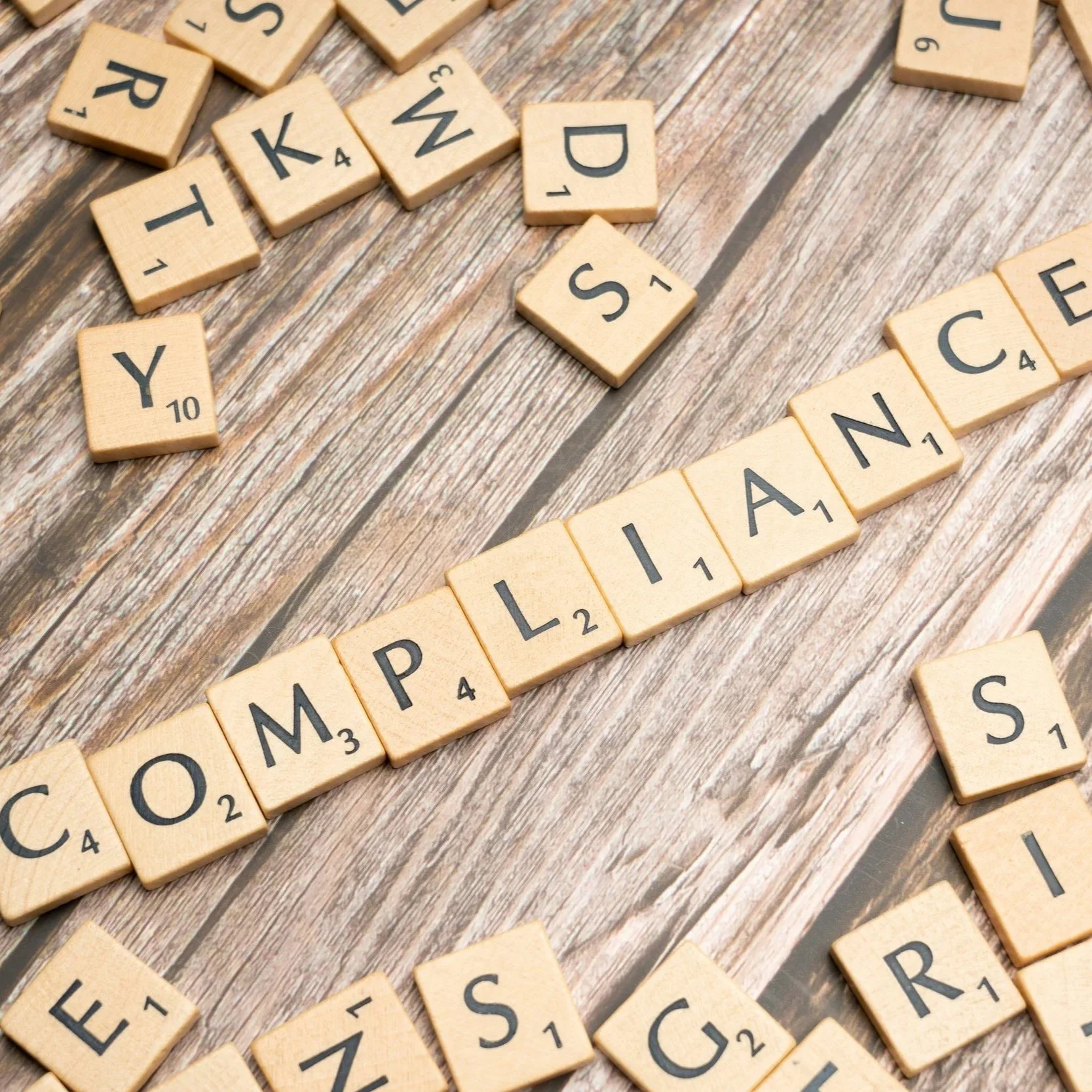 Scrabble tiles spelling out the words 'COMPLIANCE' on a wooden surface representing Colorado Candle Studio's fully insured business and venue compliant candle rentals.