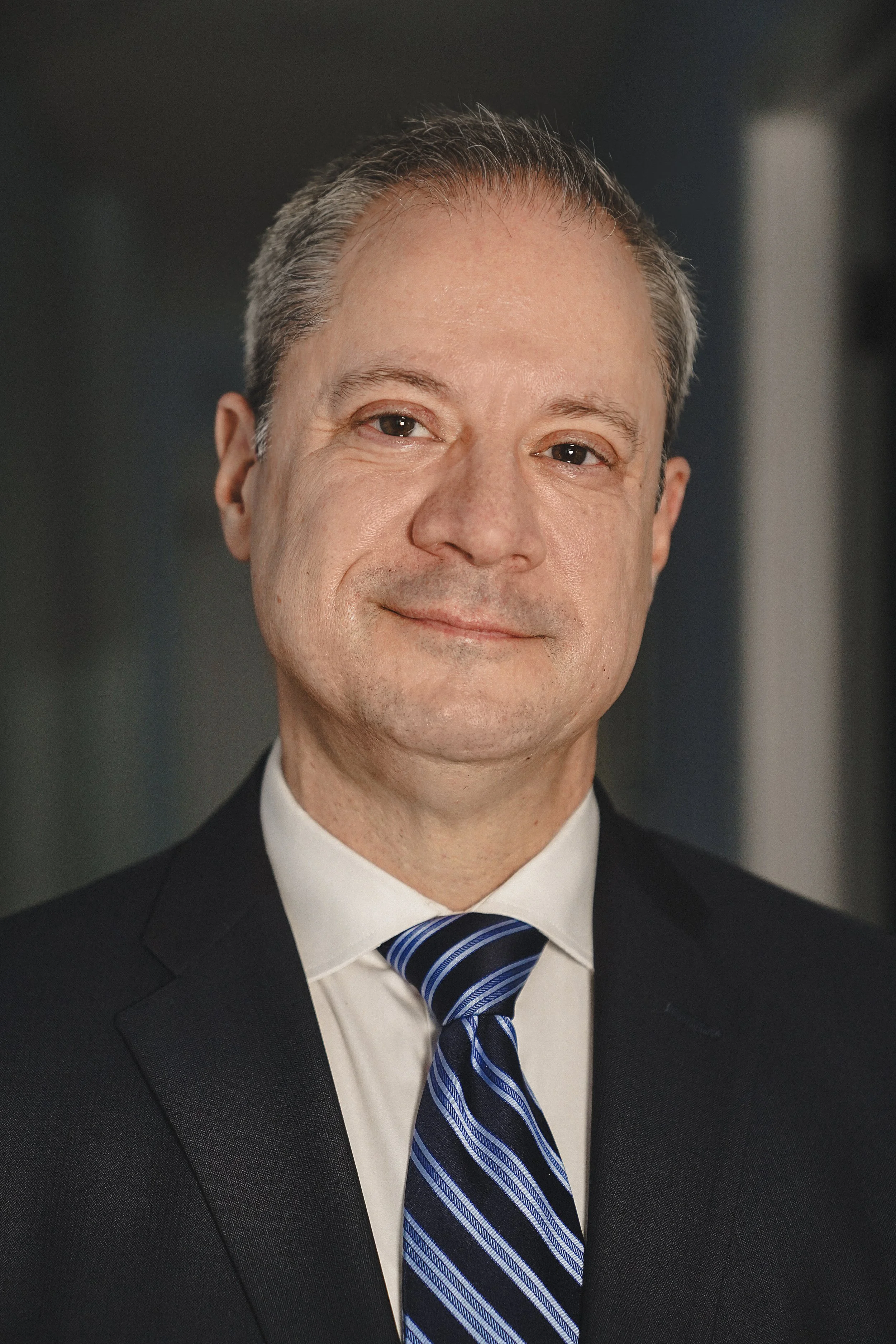 Dr. William Berger in a business suit, white shirt, and striped blue tie, smiling slightly. He has short, graying hair and is standing against a plain, dark background.