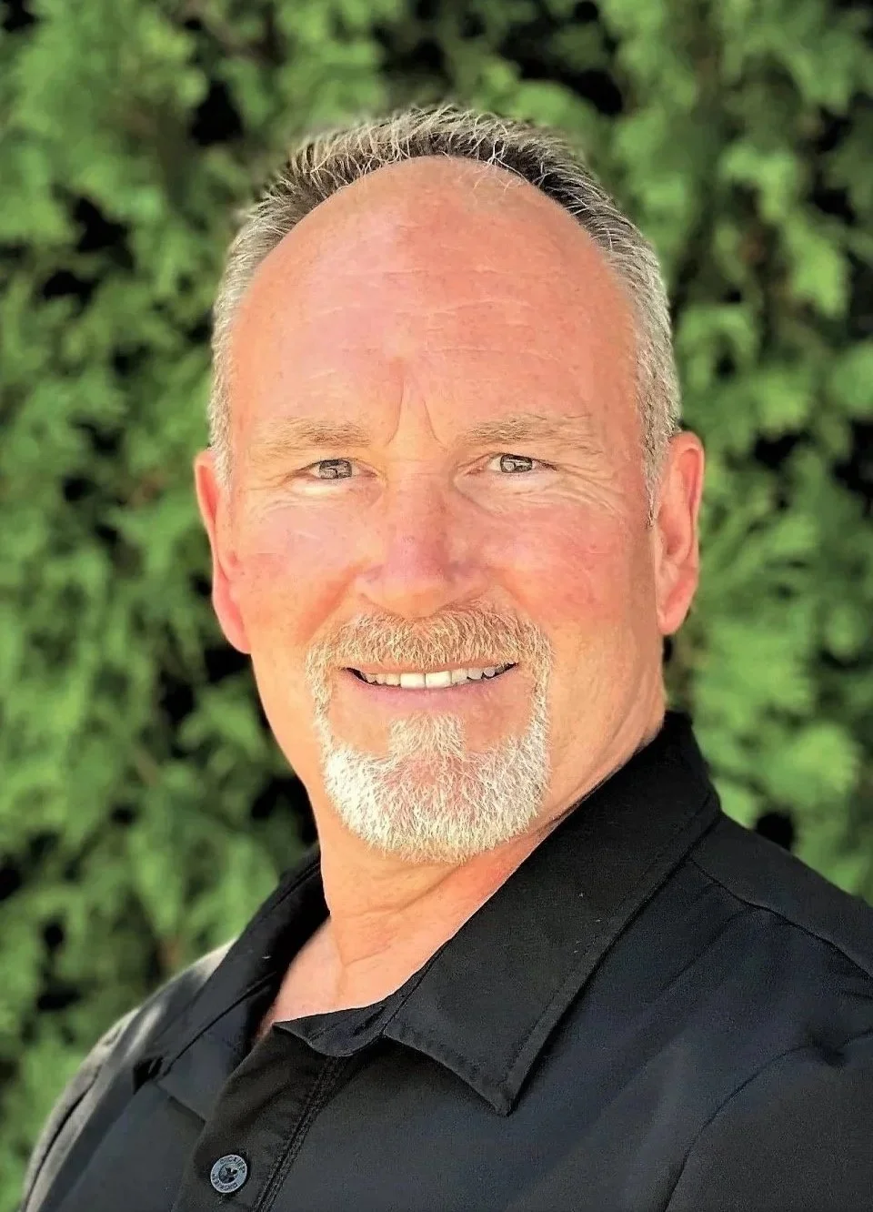 Peter Cummings with short gray hair, a goatee, and bright eyes, wearing a black shirt, standing outdoors in front of green foliage.