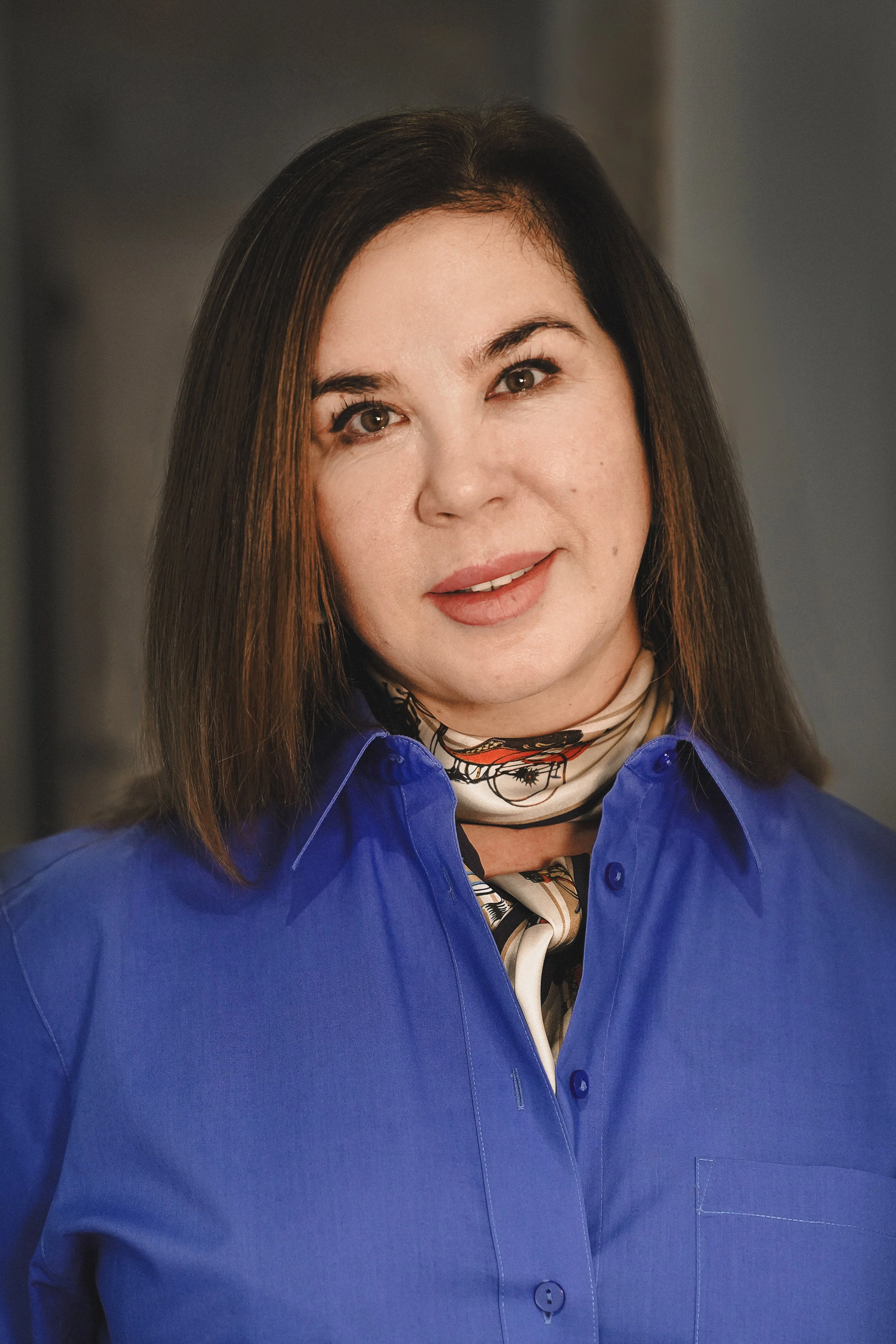 Victoria Zames with brown hair and brown eyes wearing a bright blue button-up shirt, a patterned scarf around her neck, and a floral top underneath, smiling softly.