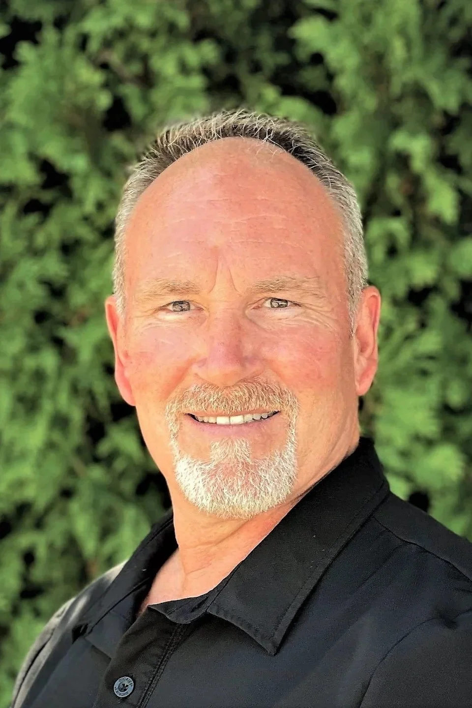 Portrait of Petter Cummings with a beard and mustache, wearing a black shirt, standing outdoors with green foliage in the background.