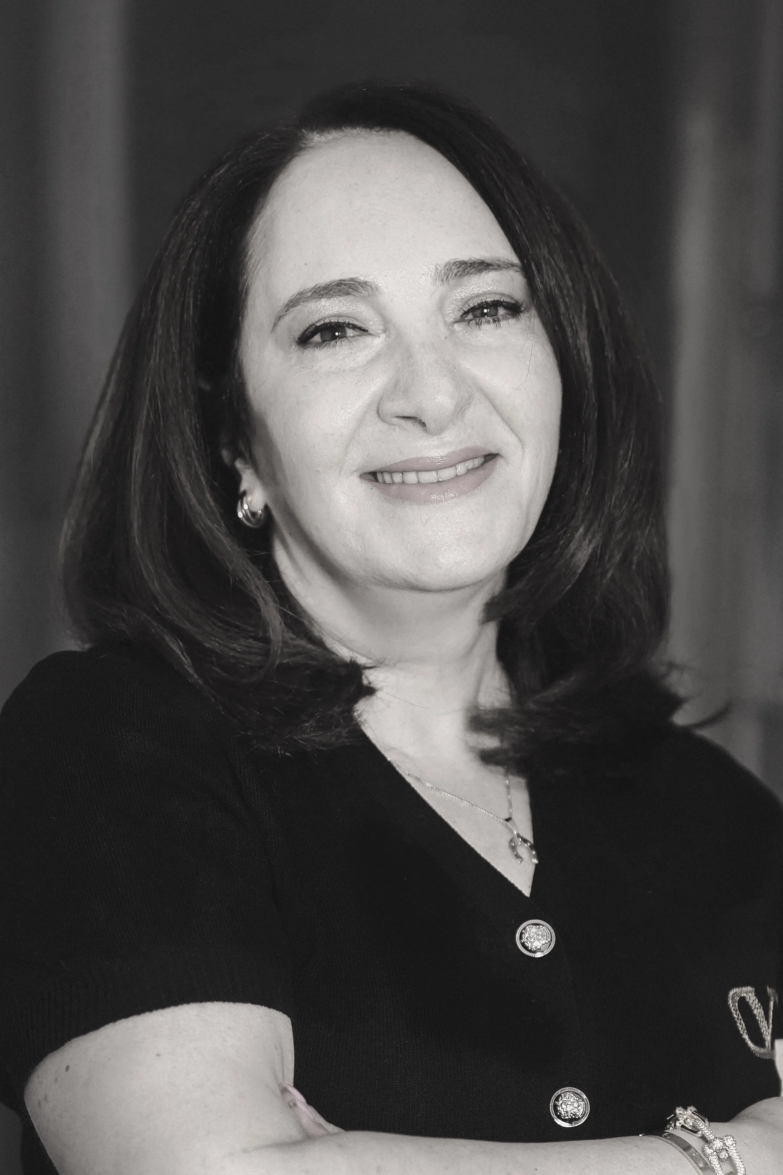 A black-and-white photo of Irina Stainov with shoulder-length dark hair, wearing earrings, a necklace, and jewelry on her wrist, smiling softly at the camera.