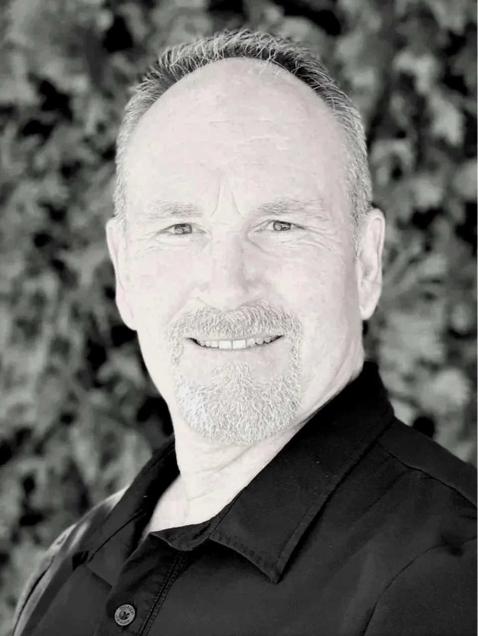 A black and white photo of Peter Cummings with short, combed back hair, facial hair, and light-colored eyes, wearing a dark collared shirt, standing outdoors with a blurred leafy background.