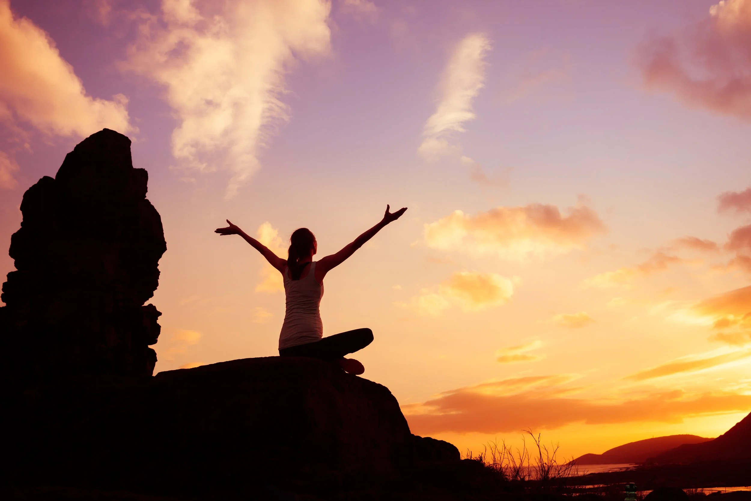 A person practicing yoga on a rock formation at sunset, with arms raised, overlooking a scenic landscape with mountains and a river under a colorful sky with clouds.