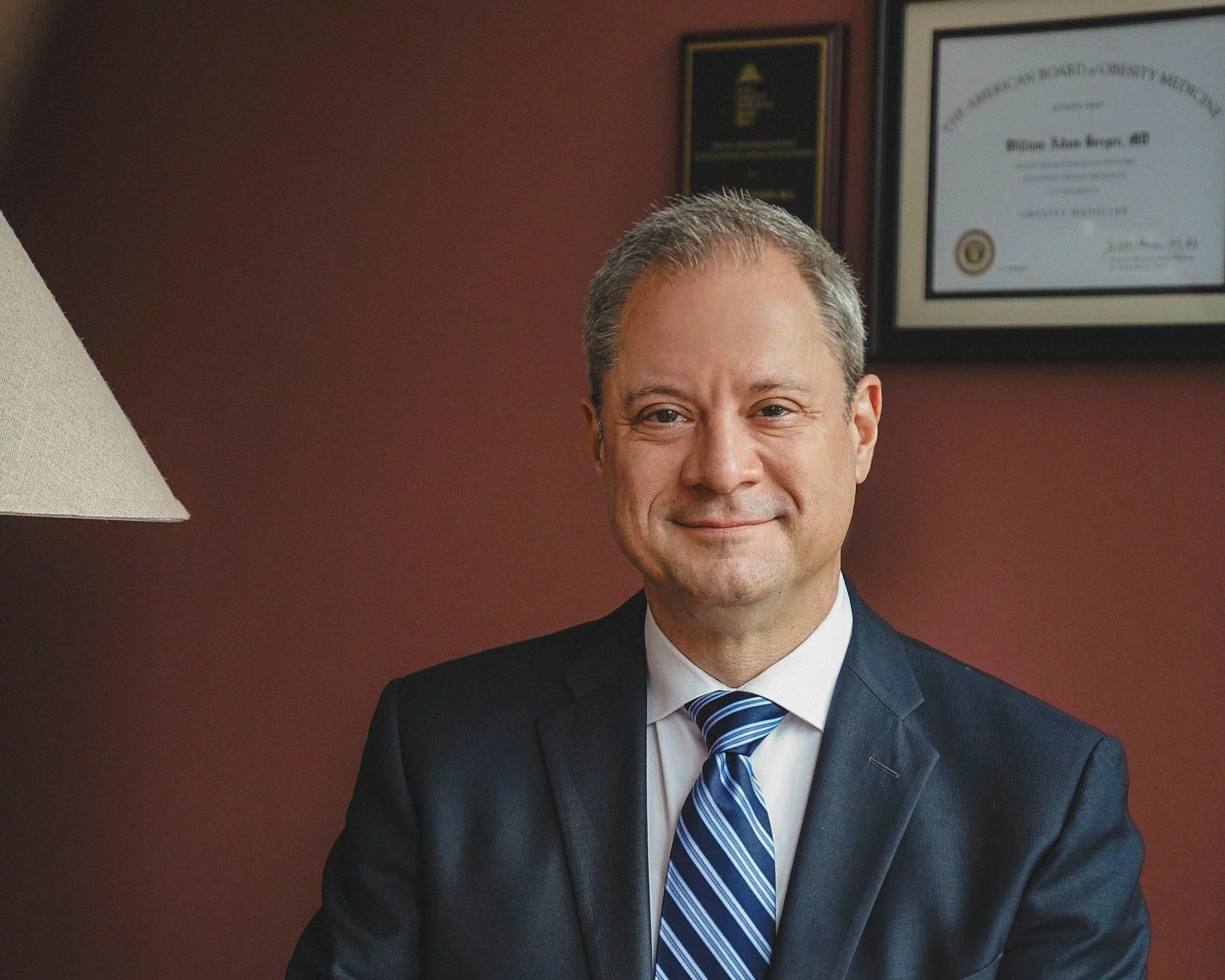 At Northeast Medical Practice Office, Close-up of Dr. William Berger in a suit, smiling, with framed certificates or awards hanging on a maroon-colored wall behind him.