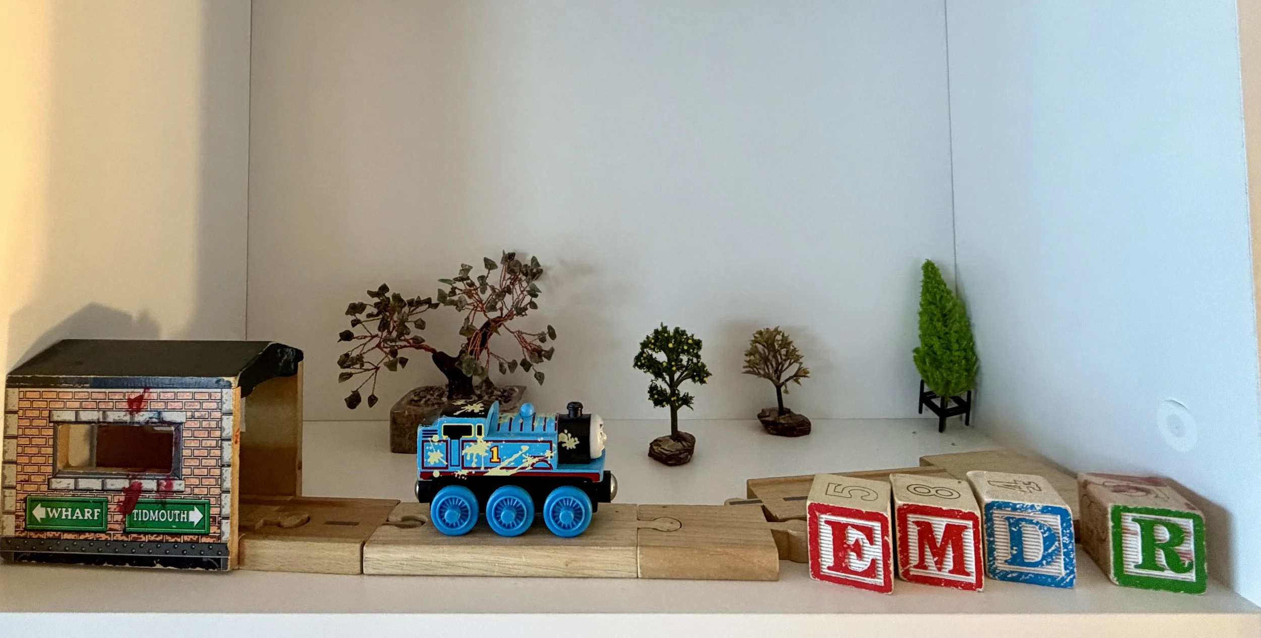 Collection of small green and brown potted trees, a toy train, a cardboard building with street signs, and alphabet blocks on a shelf.