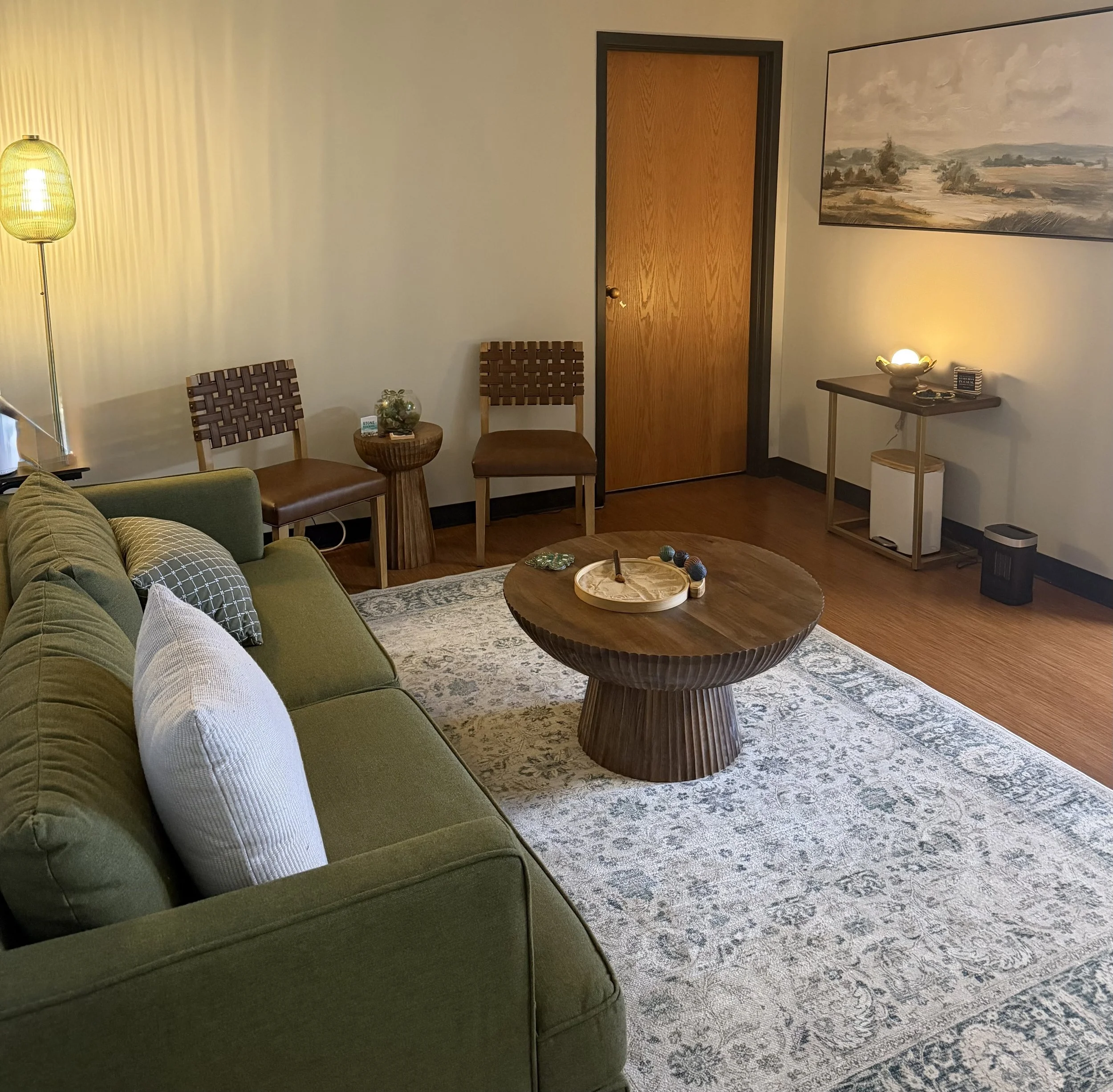 Living room with a green sofa, wooden coffee table, and decorative pillows. A patterned area rug covers the floor. There are chairs, a small side table with a plant, and a wooden door. The walls are decorated with a landscape painting and a lamp provides ambient lighting.