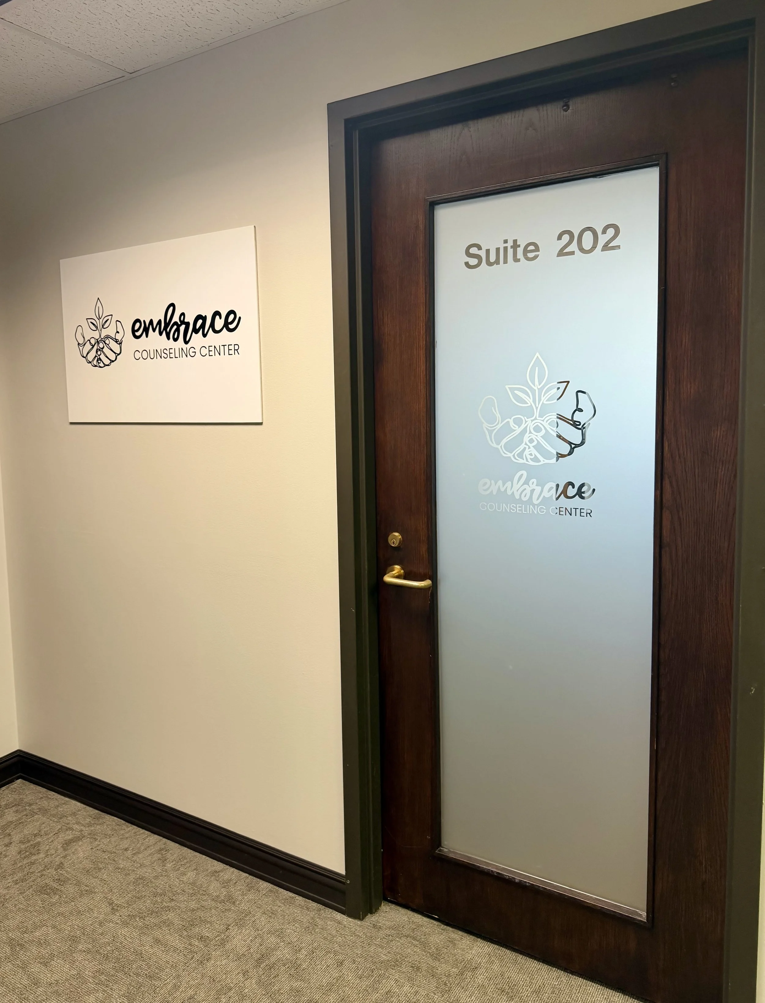 Sign on the ceiling reads 'embrace COUNSELING CENTER'. Door with glass panel has 'Suite 202' written on it, with a logo of a hand holding a plant.