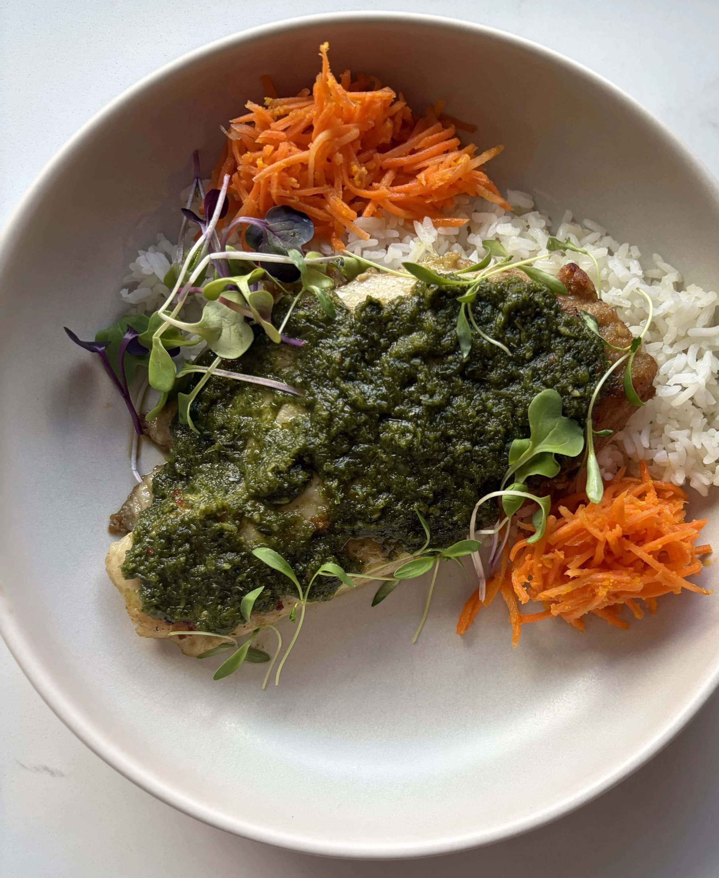 A white bowl containing rice, shredded carrots, microgreens, and a fish fillet topped with green pesto sauce.