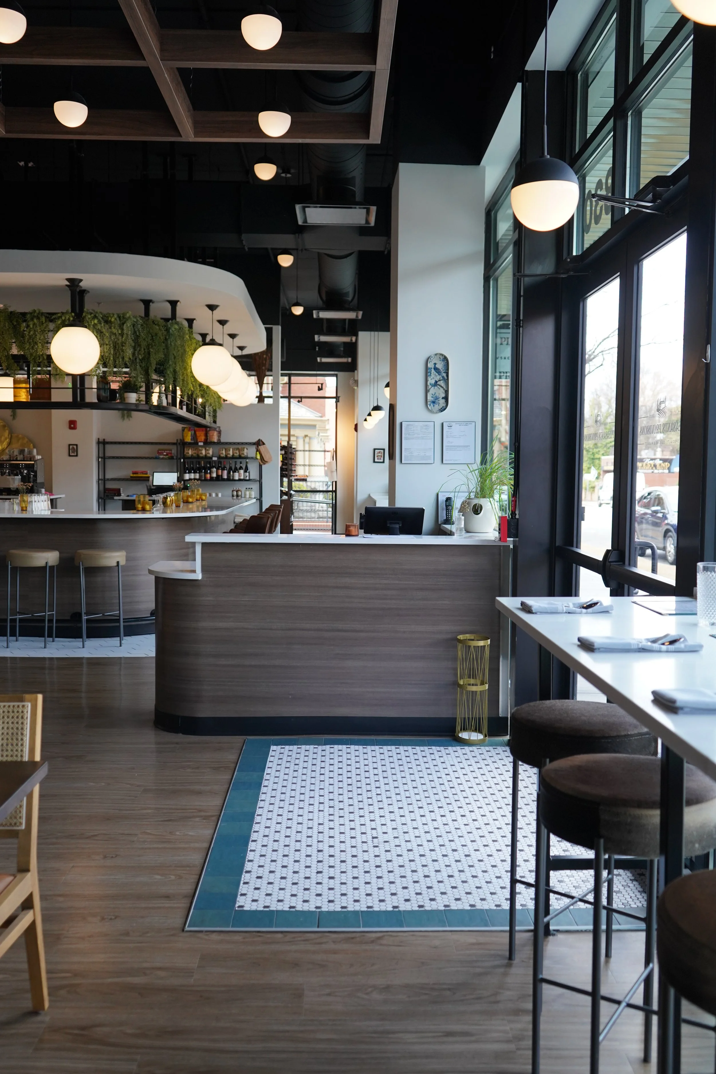 Interior of a modern café or coffee shop with white and wooden accents, hanging round white lights, a counter with bottles and shelves, a small black desk, a window with natural light, and tables with place settings.