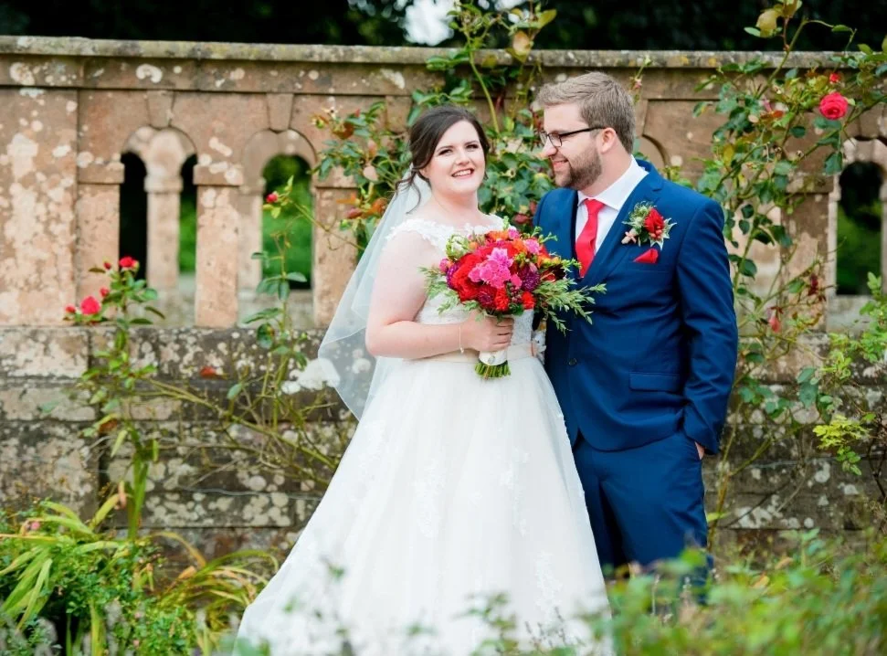A bride and groom standing outdoors in front of a stone fence with arched openings and blooming roses, smiling at each other. The bride is holding a colorful bouquet and wearing a white wedding dress with a veil, while the groom is dressed in a blue suit with a red tie and boutonniere.