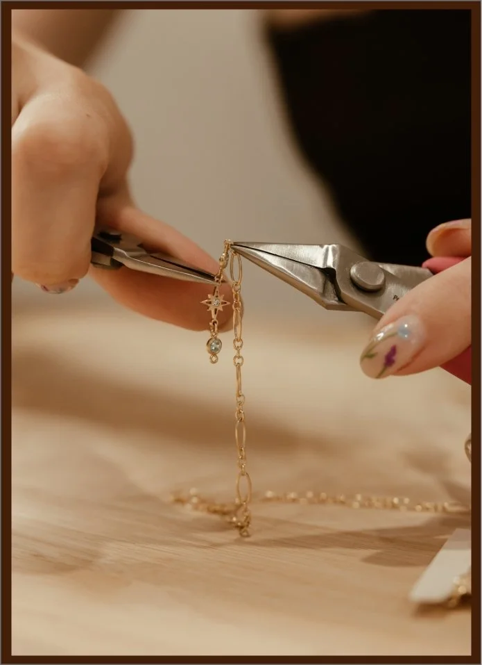 A person is using jewelry pliers to attach a small charm to a gold chain bracelet. The person's nails are decorated with floral nail art, and the background appears blurred with a wooden surface.