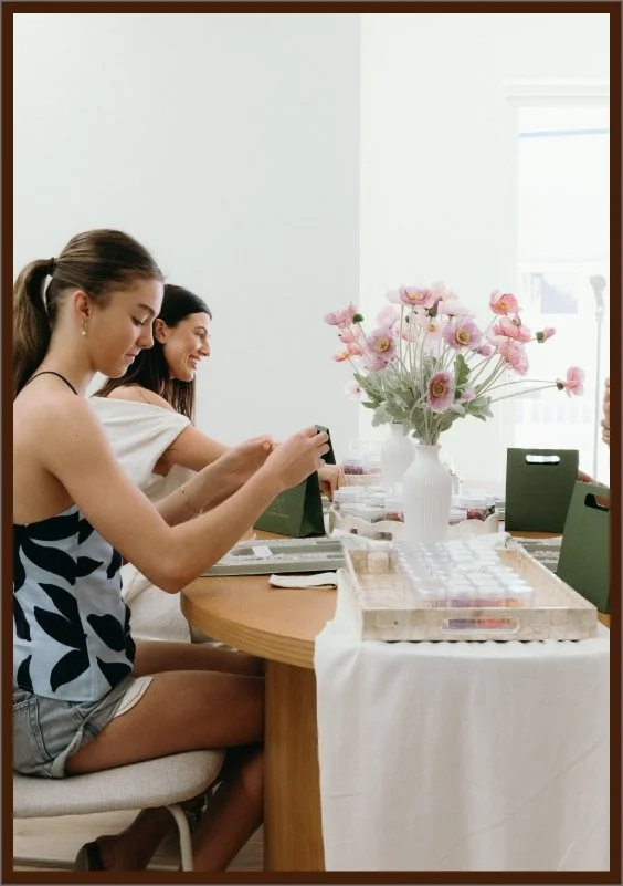 Two women sitting at a dining table with a vase of pink flowers, one woman is taking a photo of her phone, and scrapbook or craft supplies are on the table.