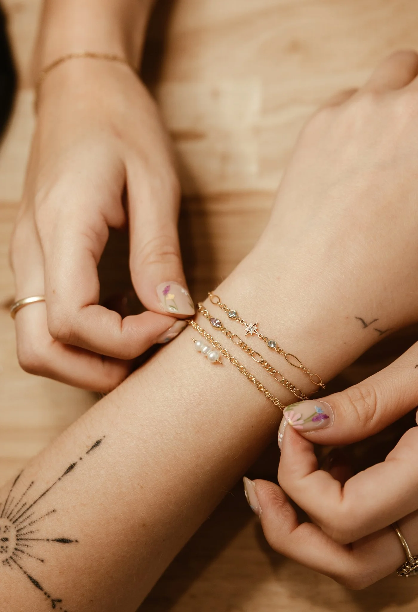 Close-up of a woman's hand and wrist adorned with gold jewelry, including bracelets and rings, with fresh manicure featuring floral nail art. The woman has small tattoos, including a dandelion tattoo on her forearm and a small bird on her wrist, and a delicate tattoo on her finger.