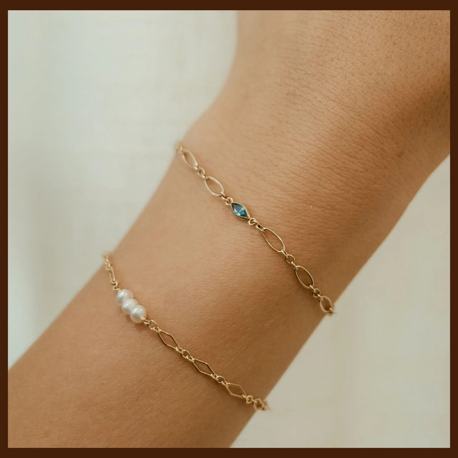Close-up of a person's wrist wearing two delicate gold chain bracelets, one with white pearls and the other with a small blue gemstone. The background is plain and neutral.