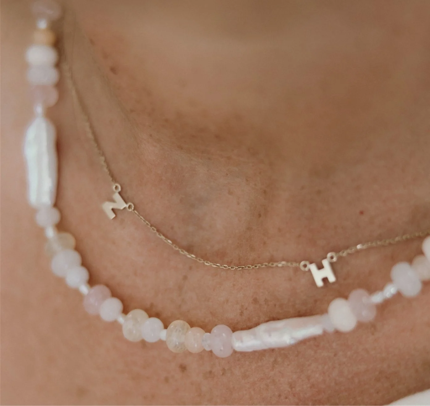 Close-up of a person's neck showing two necklaces: one with white beads and a second with a delicate chain and letter charms 'H' and 'N'.