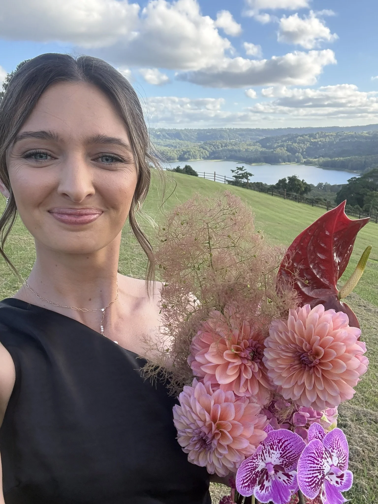 A woman with brunette hair and blue eyes smiling, holding a bouquet of pink and purple flowers, with a scenic background of a lake, green hills, trees, and partly cloudy sky.