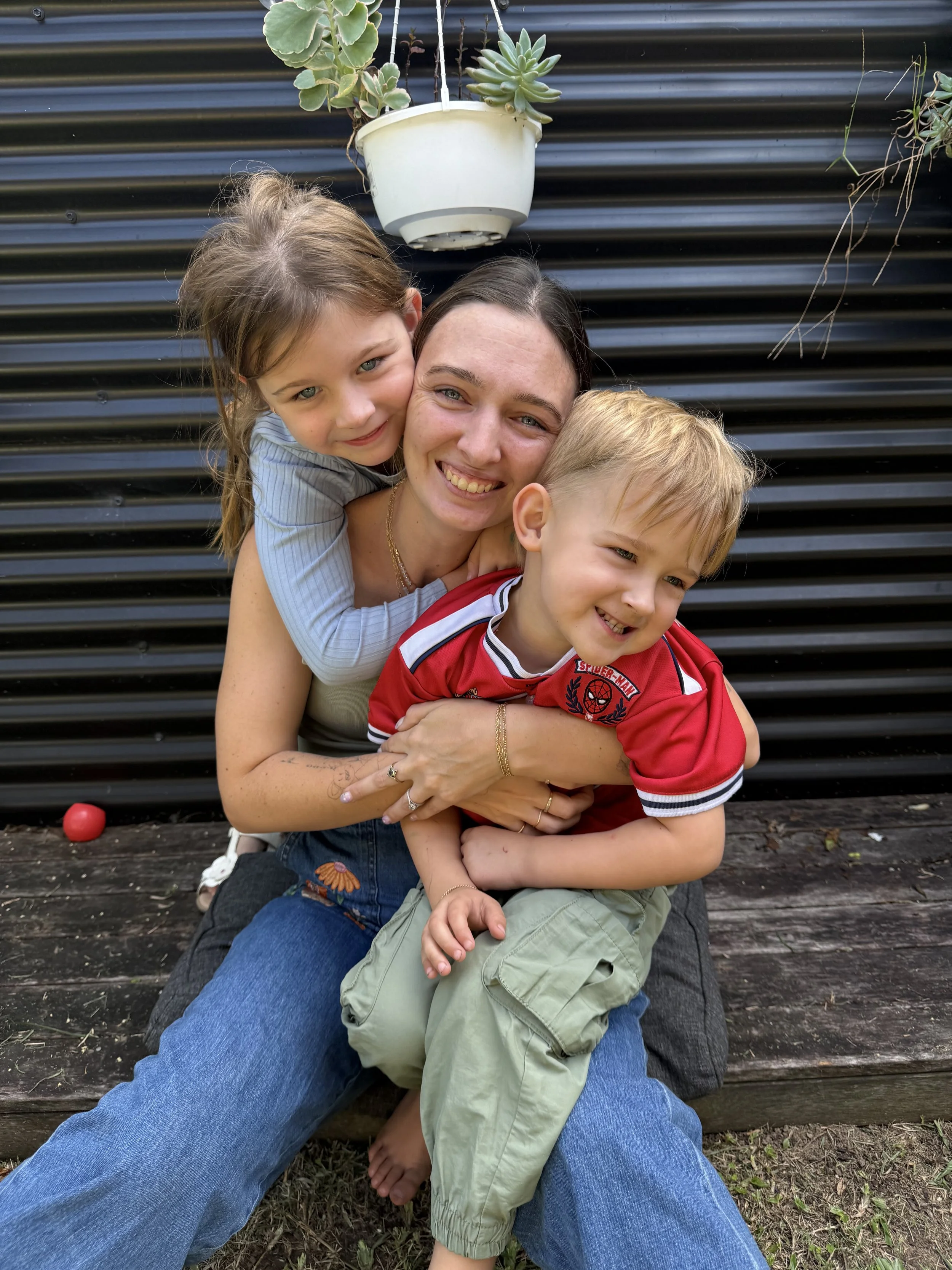 A woman and two children sitting outdoors against a black metal fence, smiling and hugging.