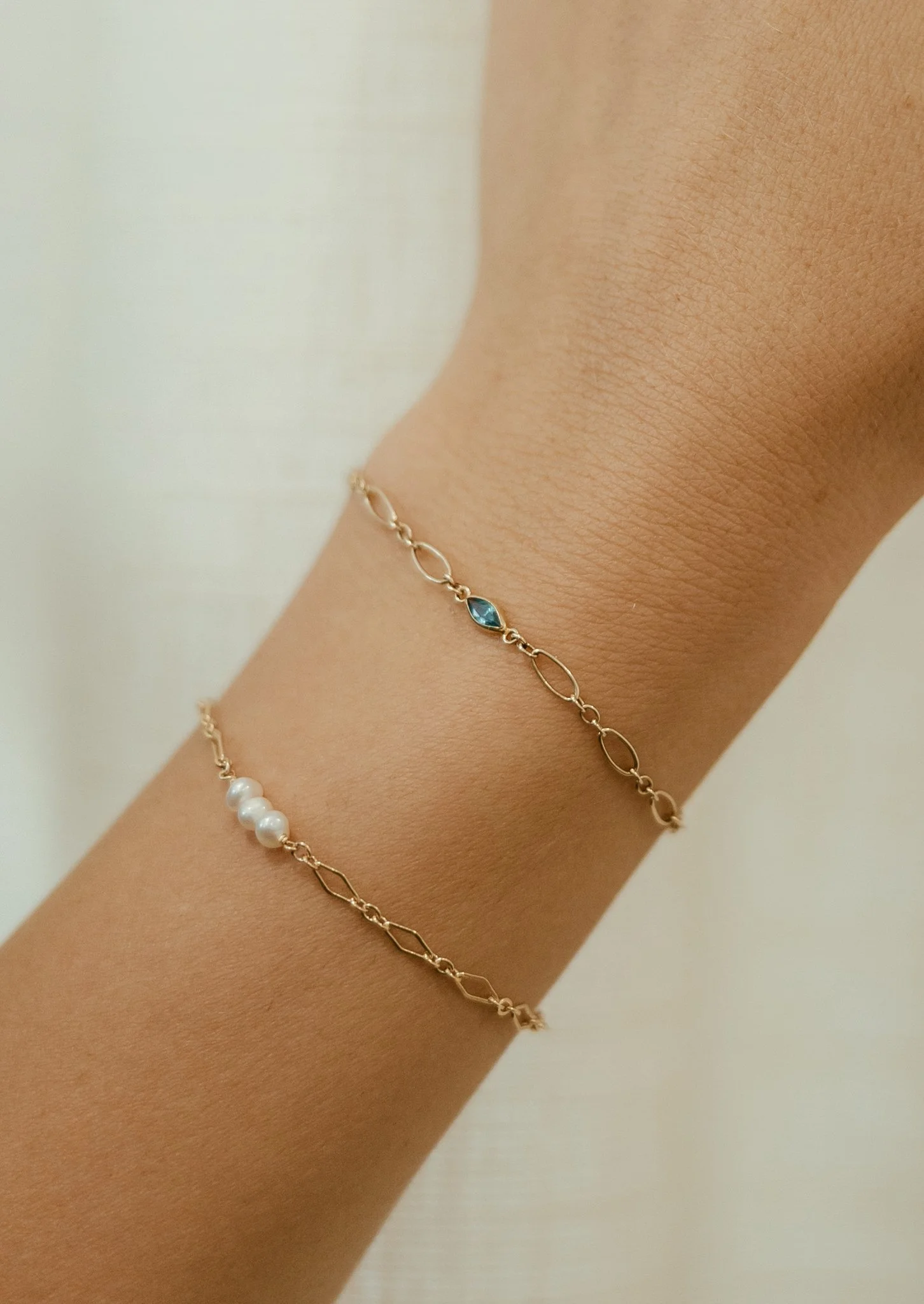 Close-up of a person's wrist with two delicate gold chain bracelets, one with white pearls and the other with blue gemstone accents.