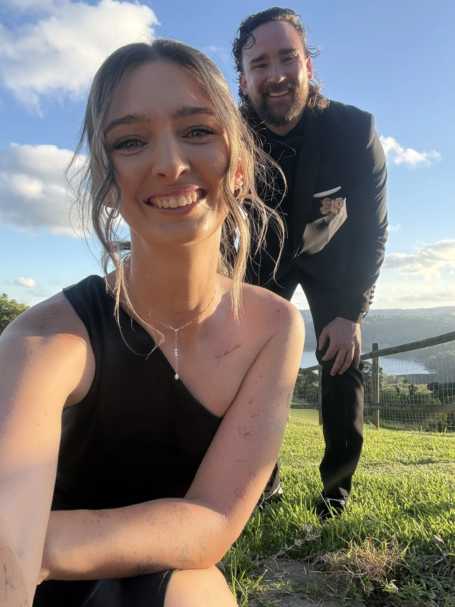 A woman with blue eyes and freckles taking a selfie outdoors, smiling with her tongue slightly out, while a bearded man in a black suit with a pink rose boutonniere stands behind her, both under a partly cloudy sky with lush green scenery in the background.