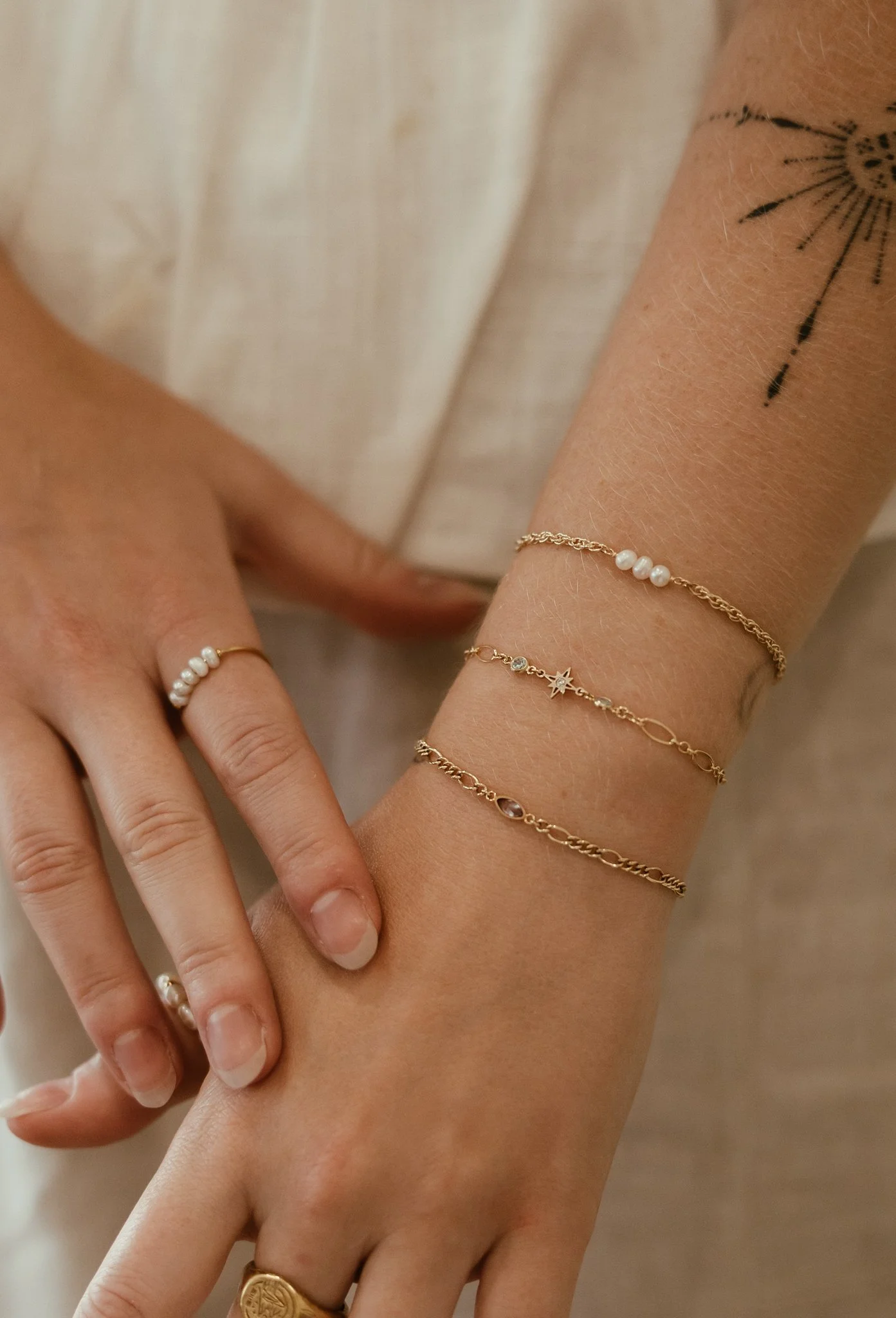 Close-up of a person's hand and wrist, adorned with gold jewelry including multiple thin bracelets and rings, and a tattoo of a partial sun or star design on the forearm.