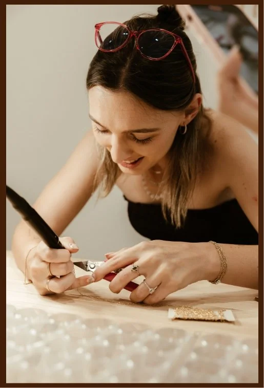A young woman with long, light brown hair, wearing a black strapless top and red glasses on her head, is smiling and leaning over a table, writing or drawing with a black pen on a piece of paper. She has a bracelet on her right wrist and rings on her fingers.
