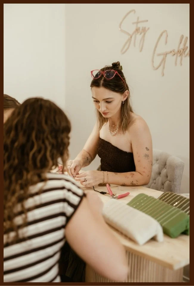 A woman with glasses on her head, tattoos, and loose hair working with a customer at a nail salon. The customer has curly hair and is wearing a striped shirt. Nail polish color samples are on the table. The background has the words 'Stay Gorgeous' on the wall.