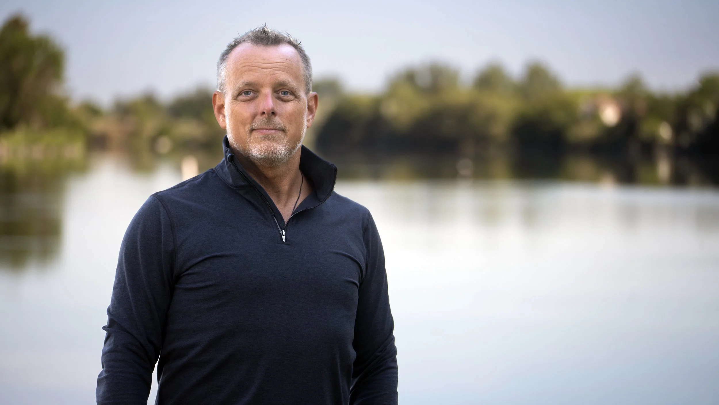 Tye Bergeson standing outdoors near a body of water, with trees and a cloudy sky in the background.