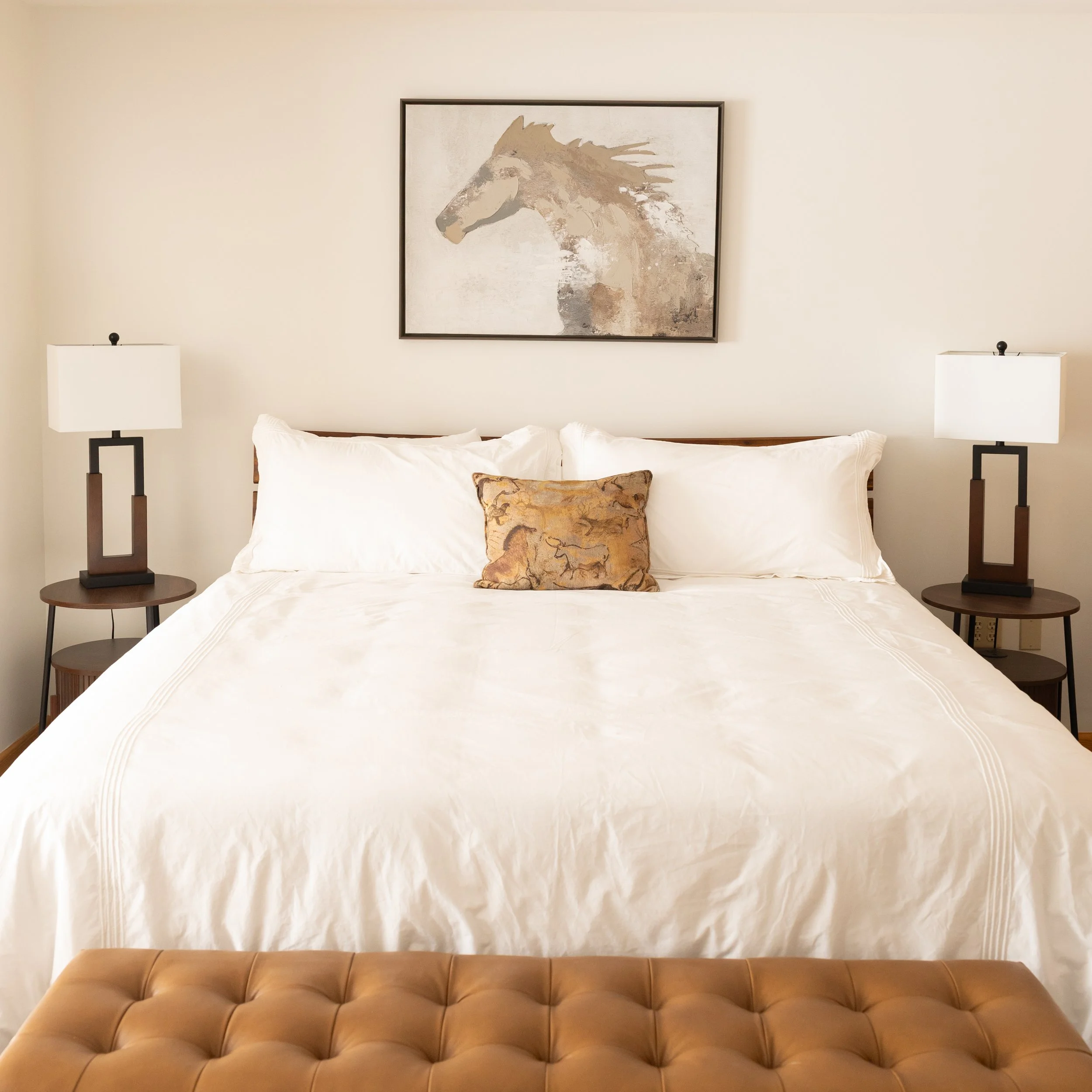 Luxury lodge and cabin rentals in Dubois, Wyoming. Dubois wedding and event venue. A bedroom with a beige headboard, white bedding, and a decorative pillow. On the wall above the bed, there is a framed abstract painting of a horse in neutral tones. 