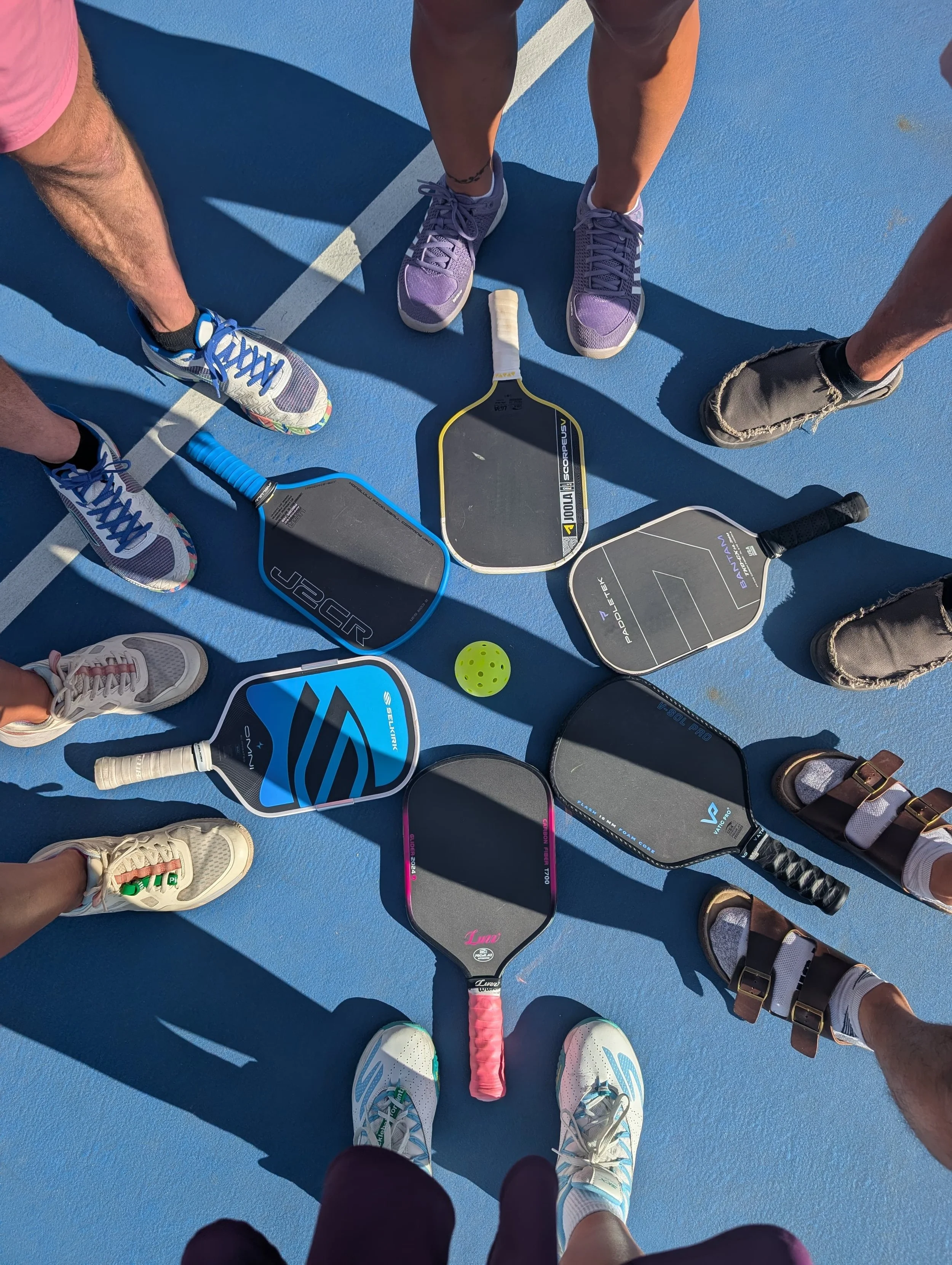Top-down view of several people standing on a pickleball court with pickleball paddles and a yellow pickleball in the center.