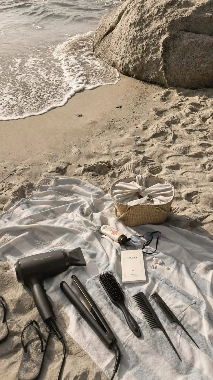 Una toalla blanca sobre la arena de playa con un secador de pelo, dos planchas de pelo, un cepillo, un peine, una crema, gafas de sol, una cesta con ropa y algunos adornos pequeños, cerca del mar con olas suaves y una roca grande en el fondo.