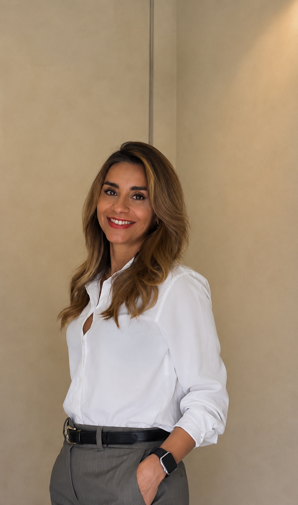 Mujer con cabello rubio, camisa blanca y pantalones grises, sonriendo y de pie con una mano en el bolsillo.
