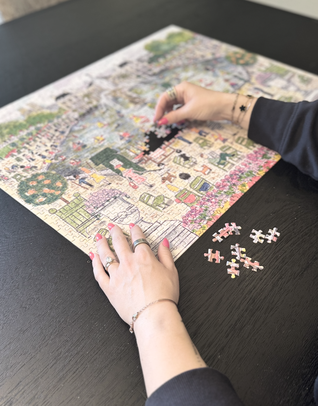 Person working on a colorful jigsaw puzzle of a scene with trees and flowers at a black table, with puzzle pieces nearby.