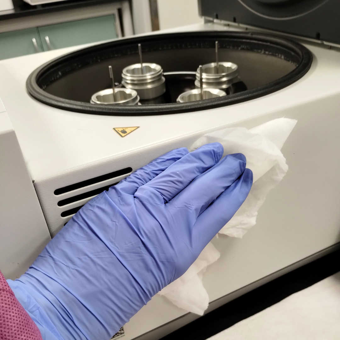 Person wearing a blue glove cleaning a laboratory centrifuge with a cloth.