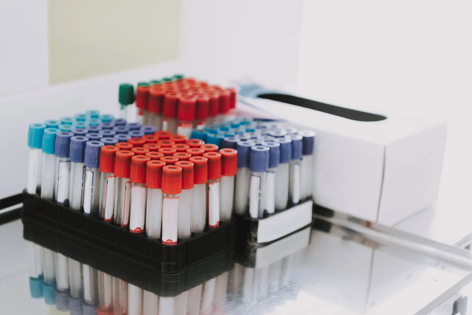 A collection of blood sample tubes with red, blue, and green caps stored in a black rack on a shiny metallic surface, with a white laboratory machine in the background.