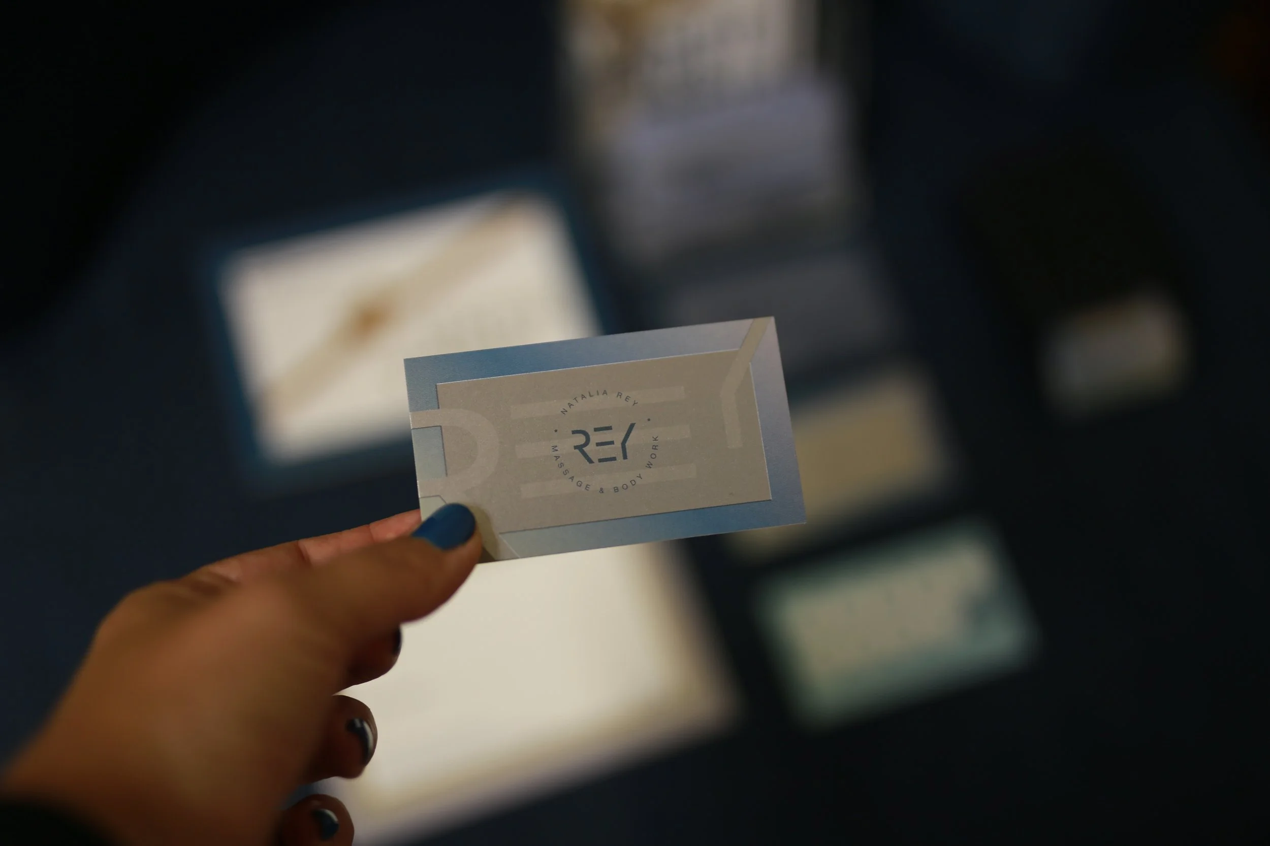 A person with blue nail polish holding a business card for 'NATALIA REY Massage & Body Work' with a blurred background.