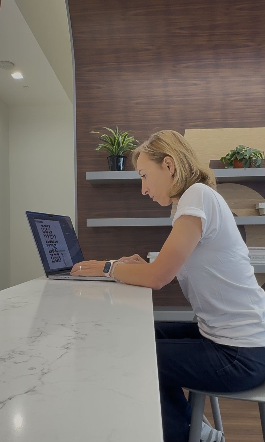 A woman with shoulder-length blonde hair sitting at a white marble table using a laptop, in a modern office with wooden wall accents and potted plants.