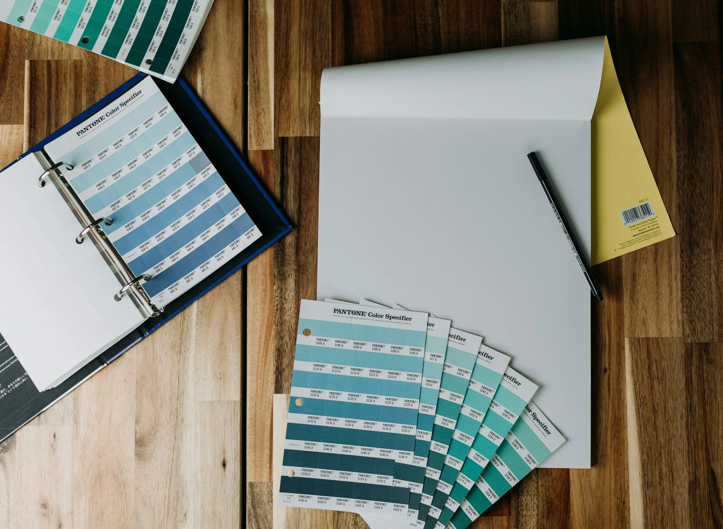 Multiple Pantone color swatch samples laid out on a wooden table, with a color match chart booklet, a book, and a pen nearby.