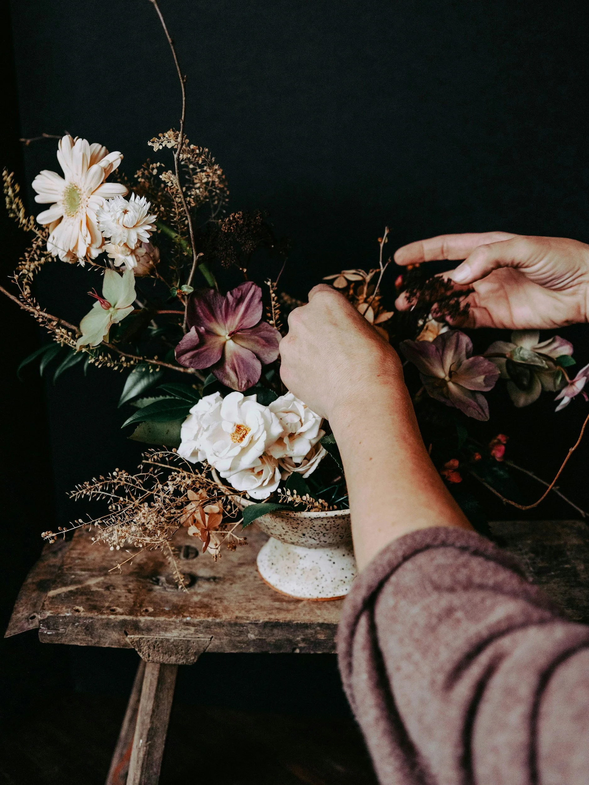 Person arranging dried flowers in a vase on a wooden table against a black background.