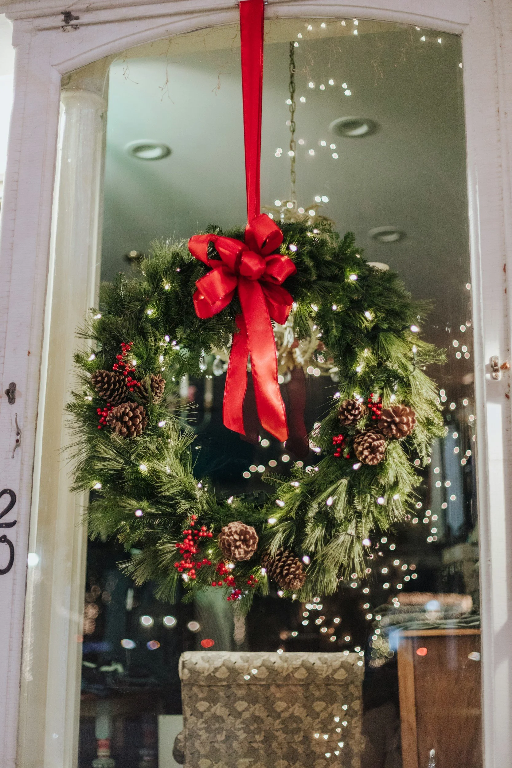A Christmas wreath with pinecones, red berries, and a red bow hanging inside a window, with Christmas lights and decorations visible through the window.