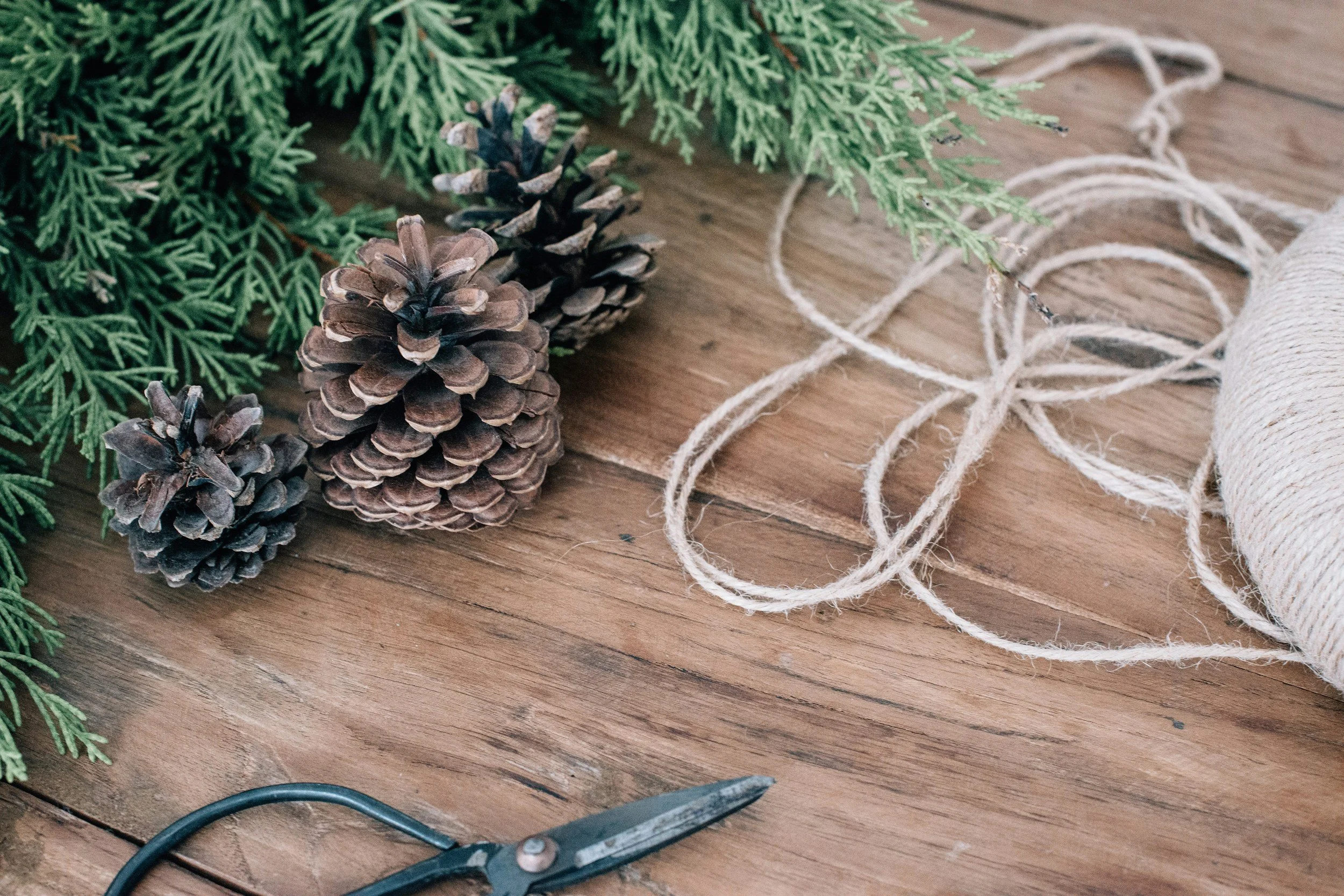 Evergreen branches, pine cones, twine, and scissors on wooden surface.
