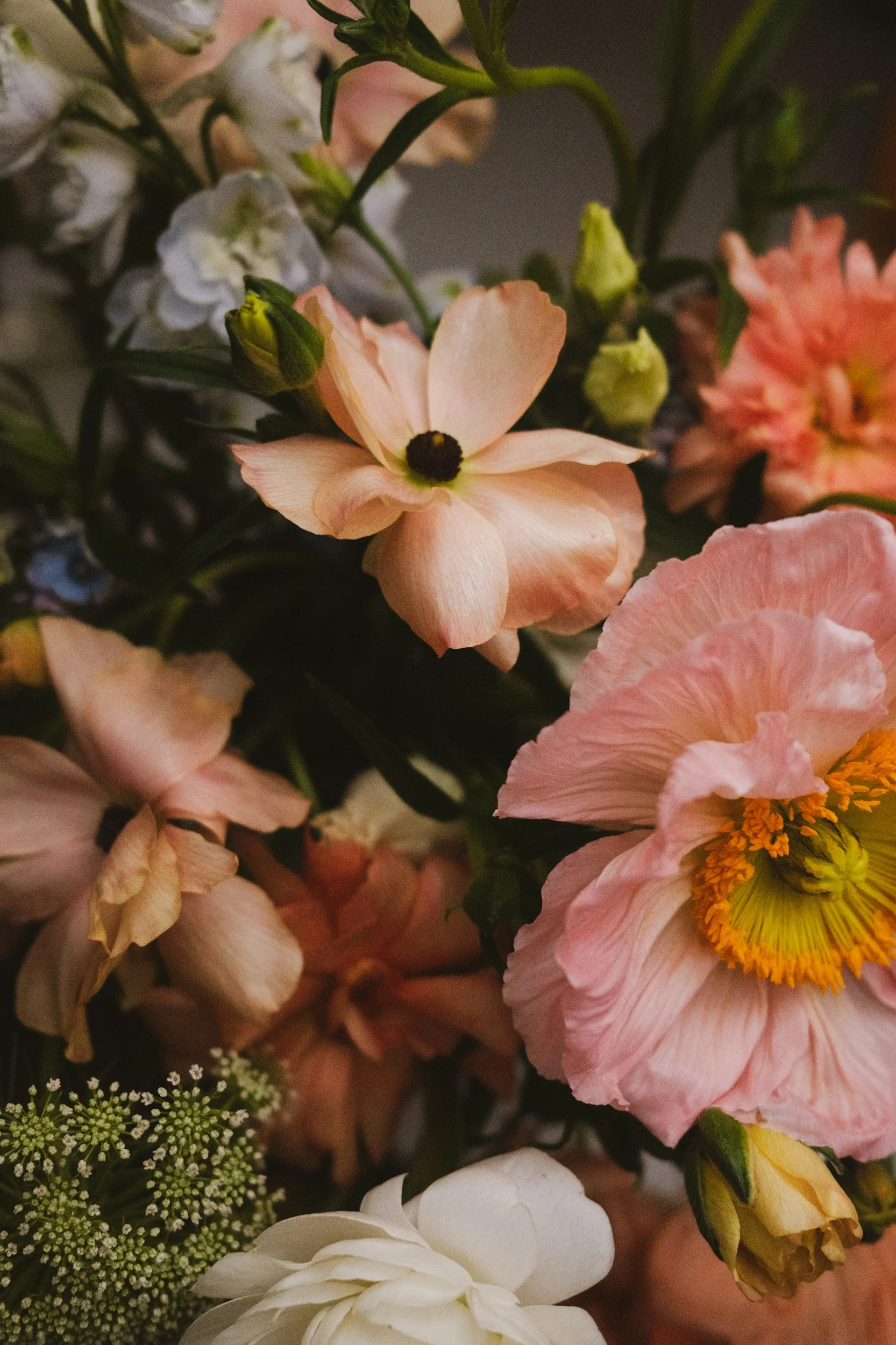 A close-up of a bouquet with pink, white, and peach flowers, including a peach-colored flower with dark center and a pink flower with yellow center, surrounded by green foliage.